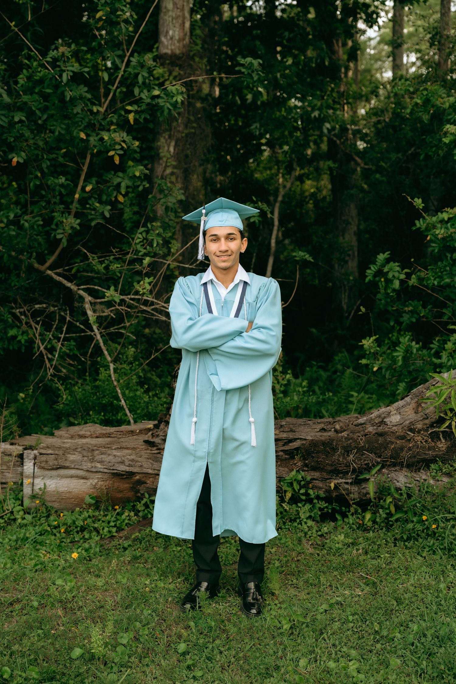 Young man in light blue graduation cap and gown stands with arms crossed outdoors in a wooded setting. Senior Photos at Colt Farms