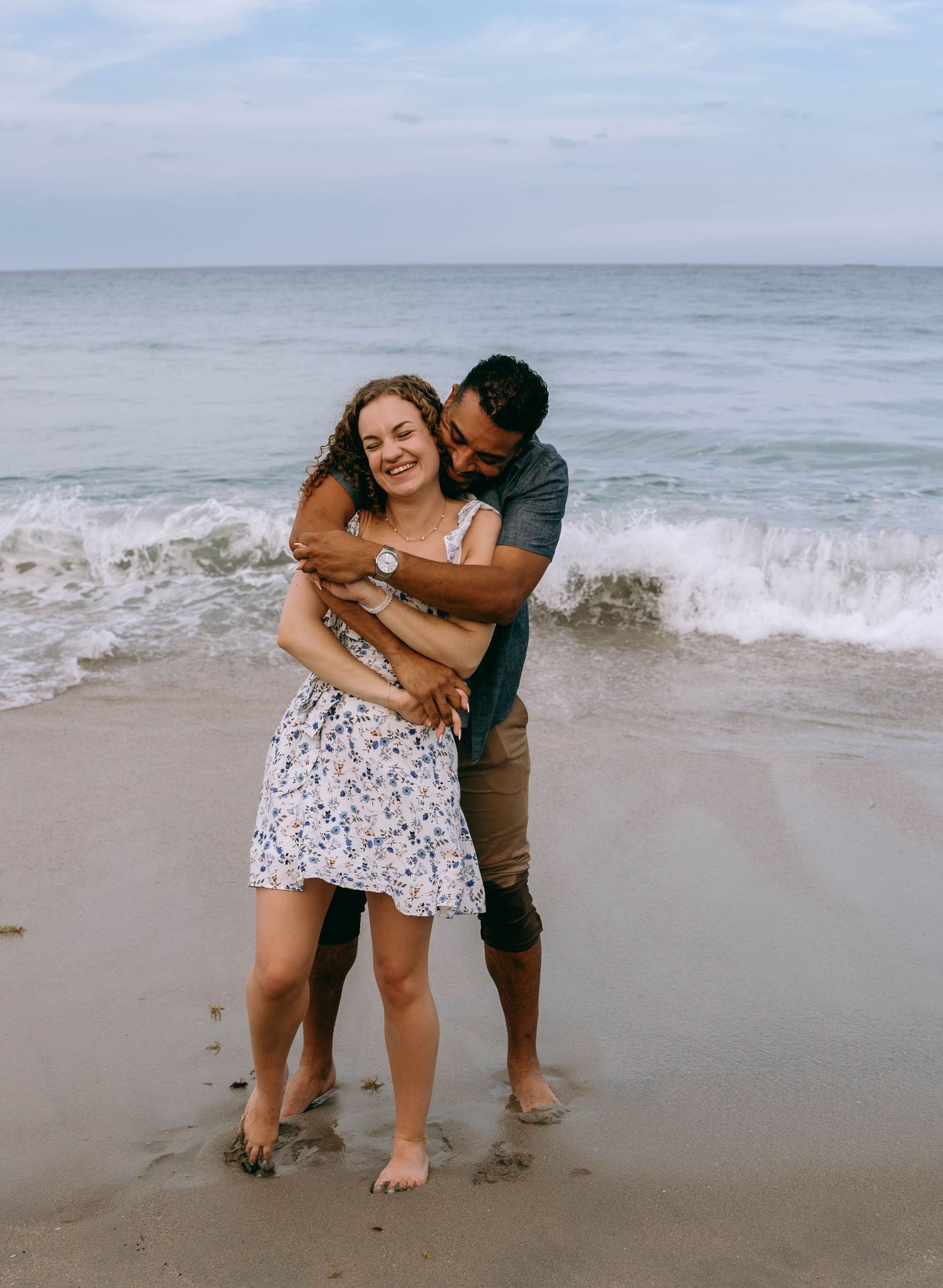 Couple embracing and laughing together on a wet sandy beach with waves crashing behind them on Worth Avenue Palm beach.
