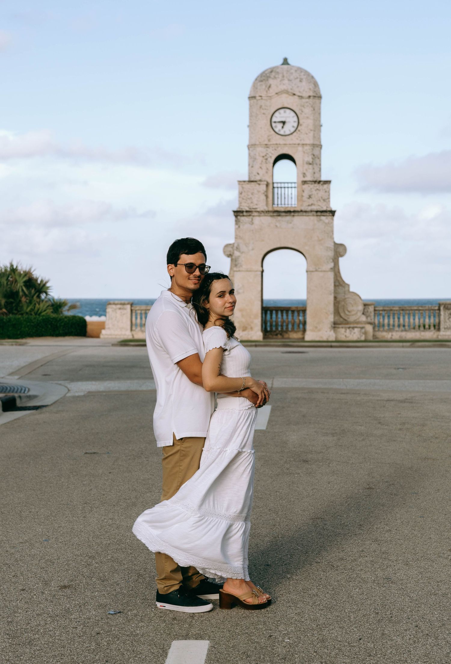 A couple in white and tan outfits pose in front of a historic clock tower near the ocean. Engagement photos on Worth Avenue in Palm Beach