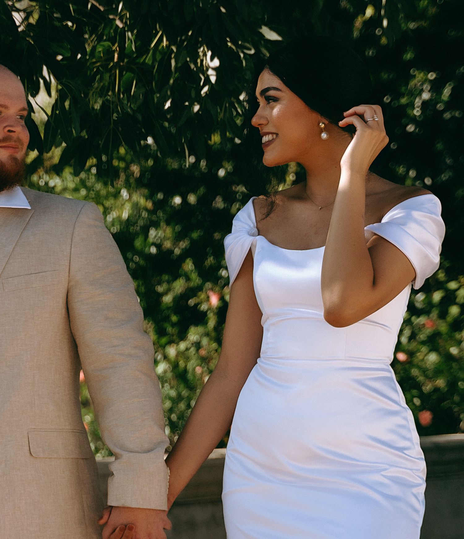 Stylish couple in white and beige posing under shaded trees.