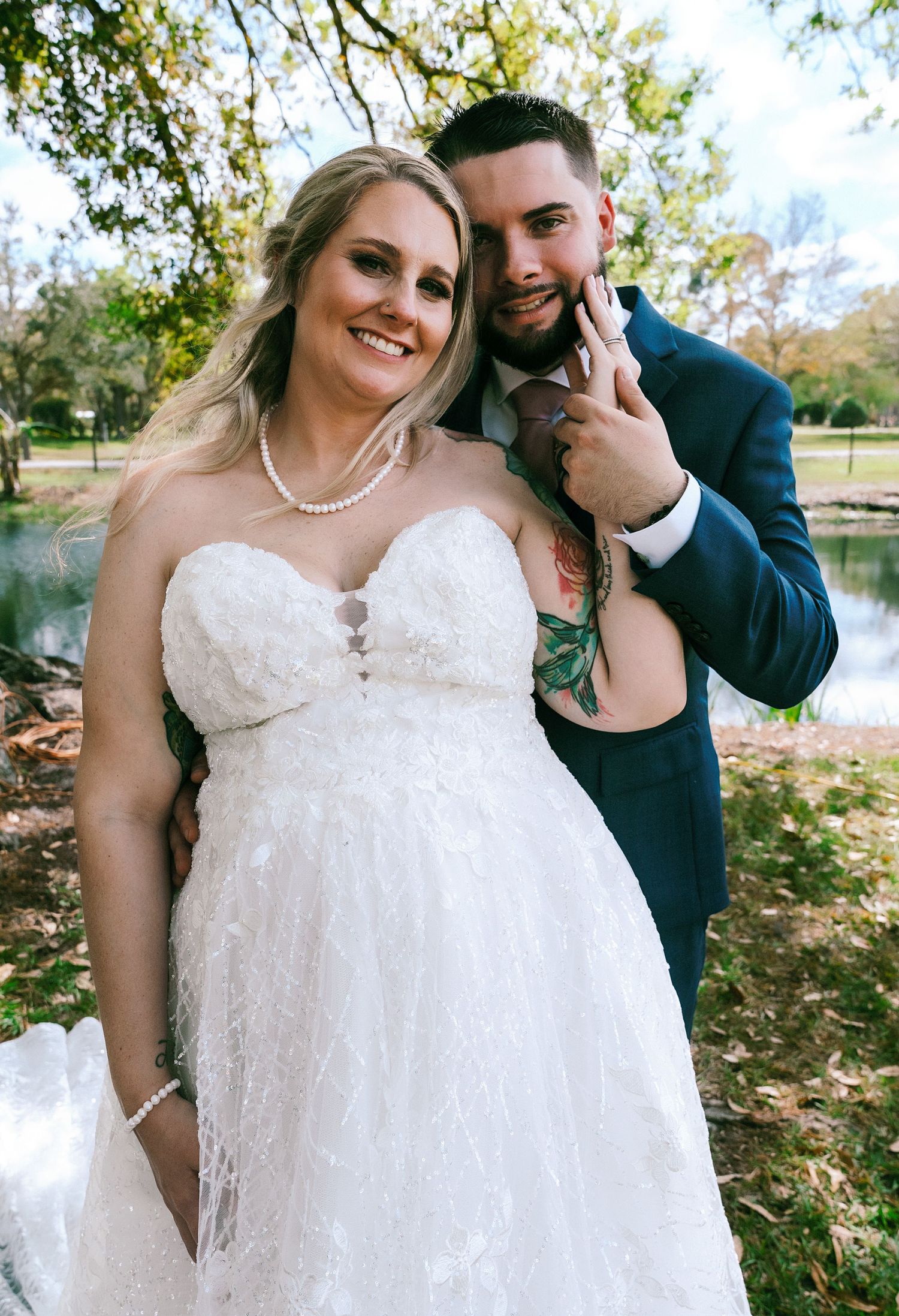 Smiling bride in strapless lace gown poses with groom in navy suit outdoors near a pond.