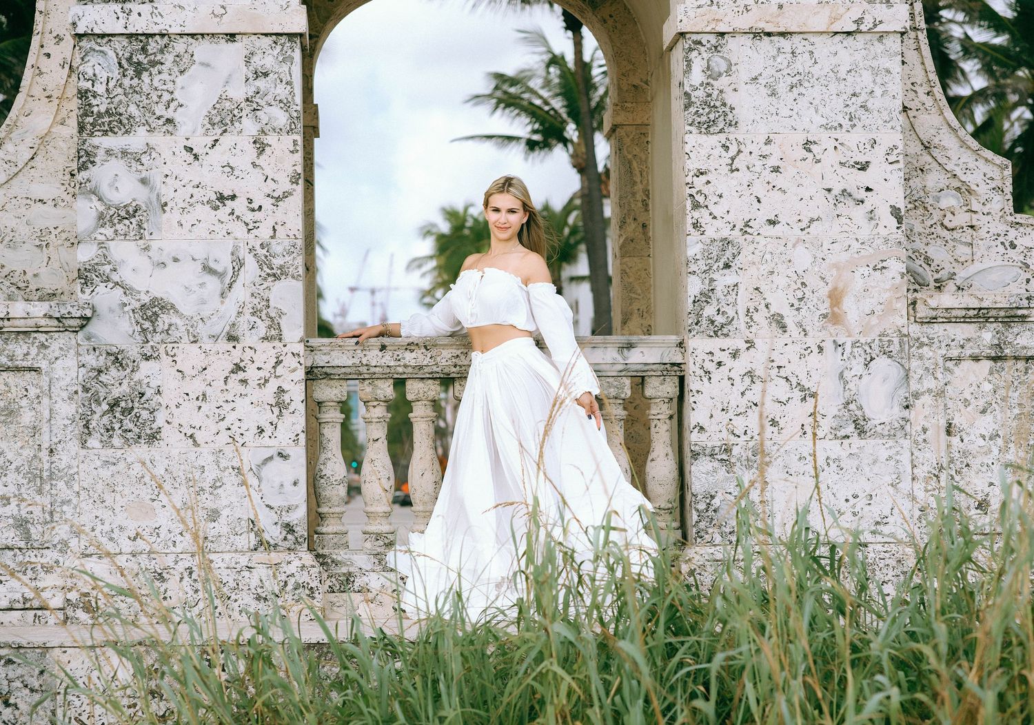 A bride in a white gown stands gracefully against a weathered stone wall surrounded by tall grass and greenery.