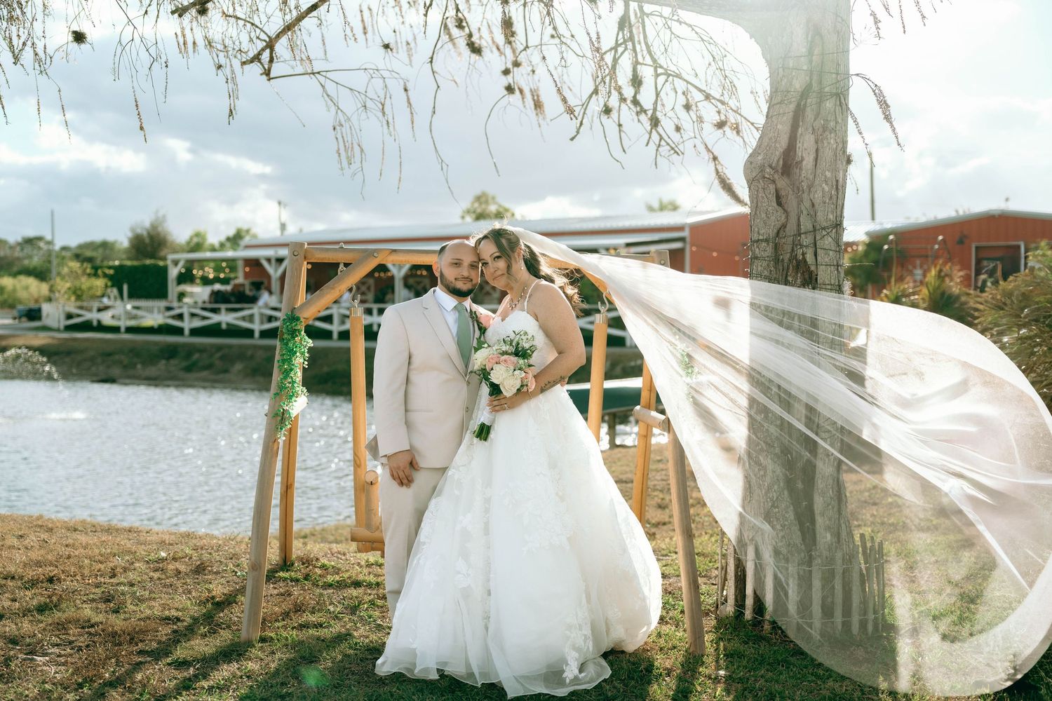 A flowing white wedding veil catches the breeze against a scenic outdoor backdrop with trees and water.