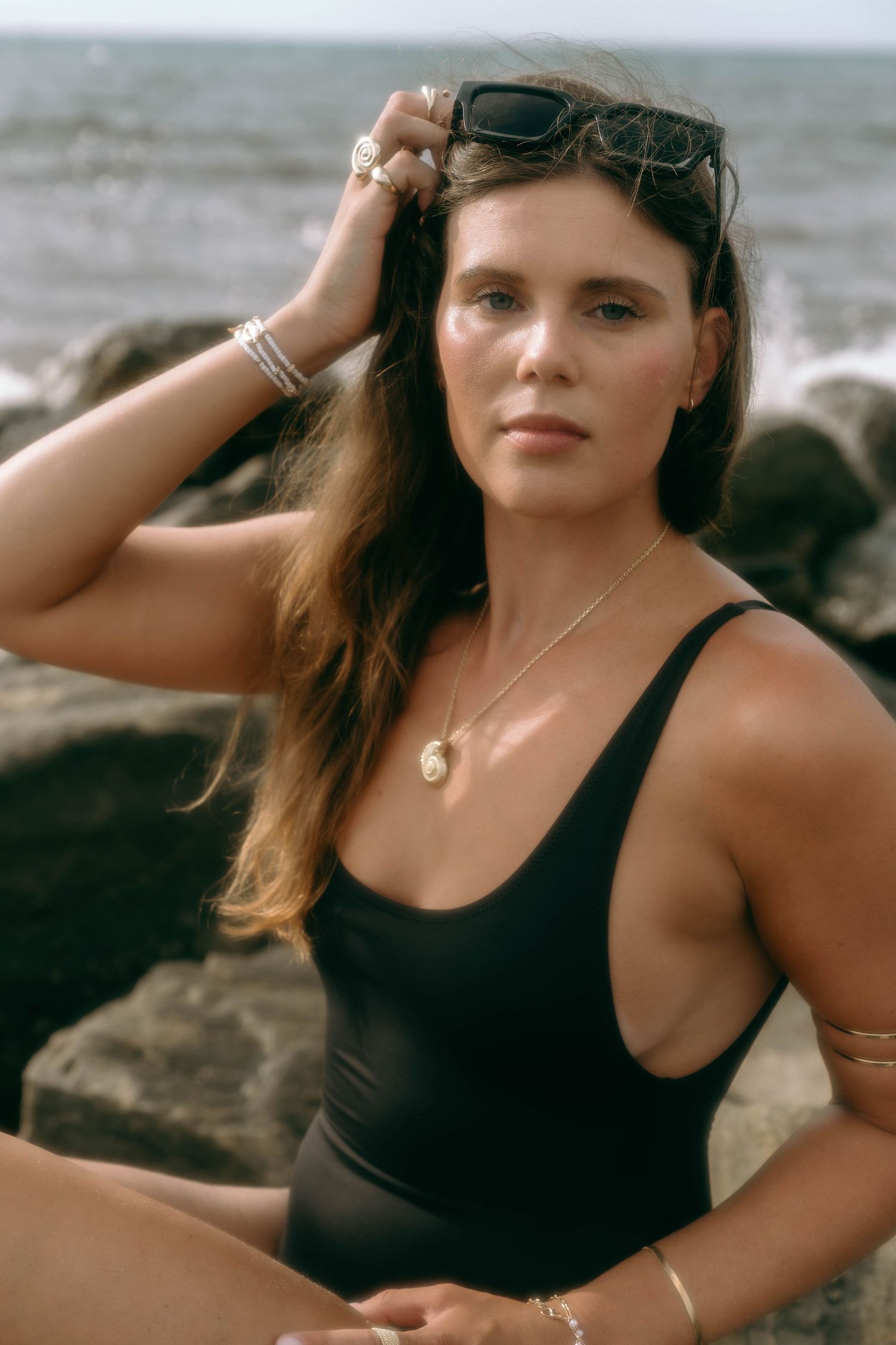 Woman in black swimsuit sitting on rocks by the ocean, wearing gold jewelry and sunglasses on her head.