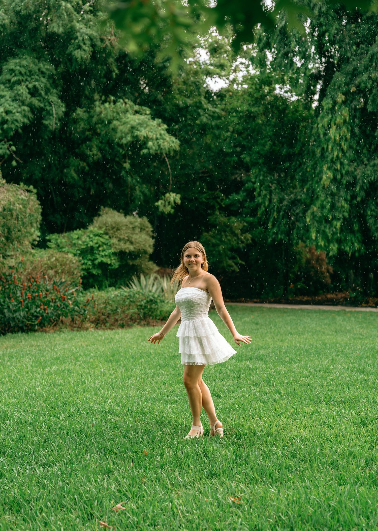 Young woman in white strapless dress twirling on green lawn surrounded by lush garden trees.