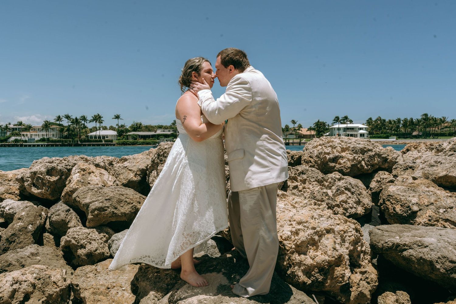 Bride and groom share a romantic moment on rocky shoreline with turquoise ocean and palm trees in the background.