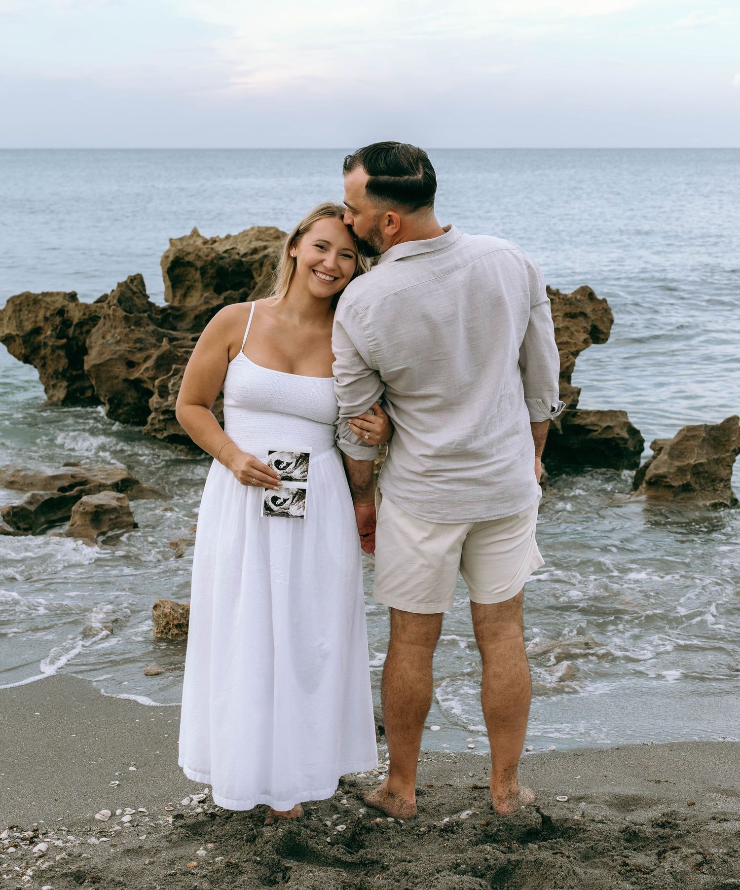 Couple sharing tender moment on rocky beach, woman in white dress holding sonogram photo.