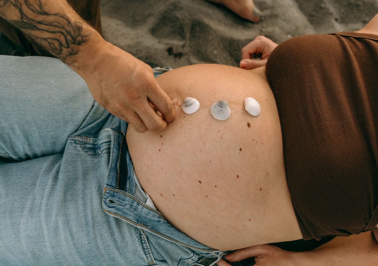 Pregnant woman lying down with small seashells placed on her bare baby bump, wearing jeans and brown top.