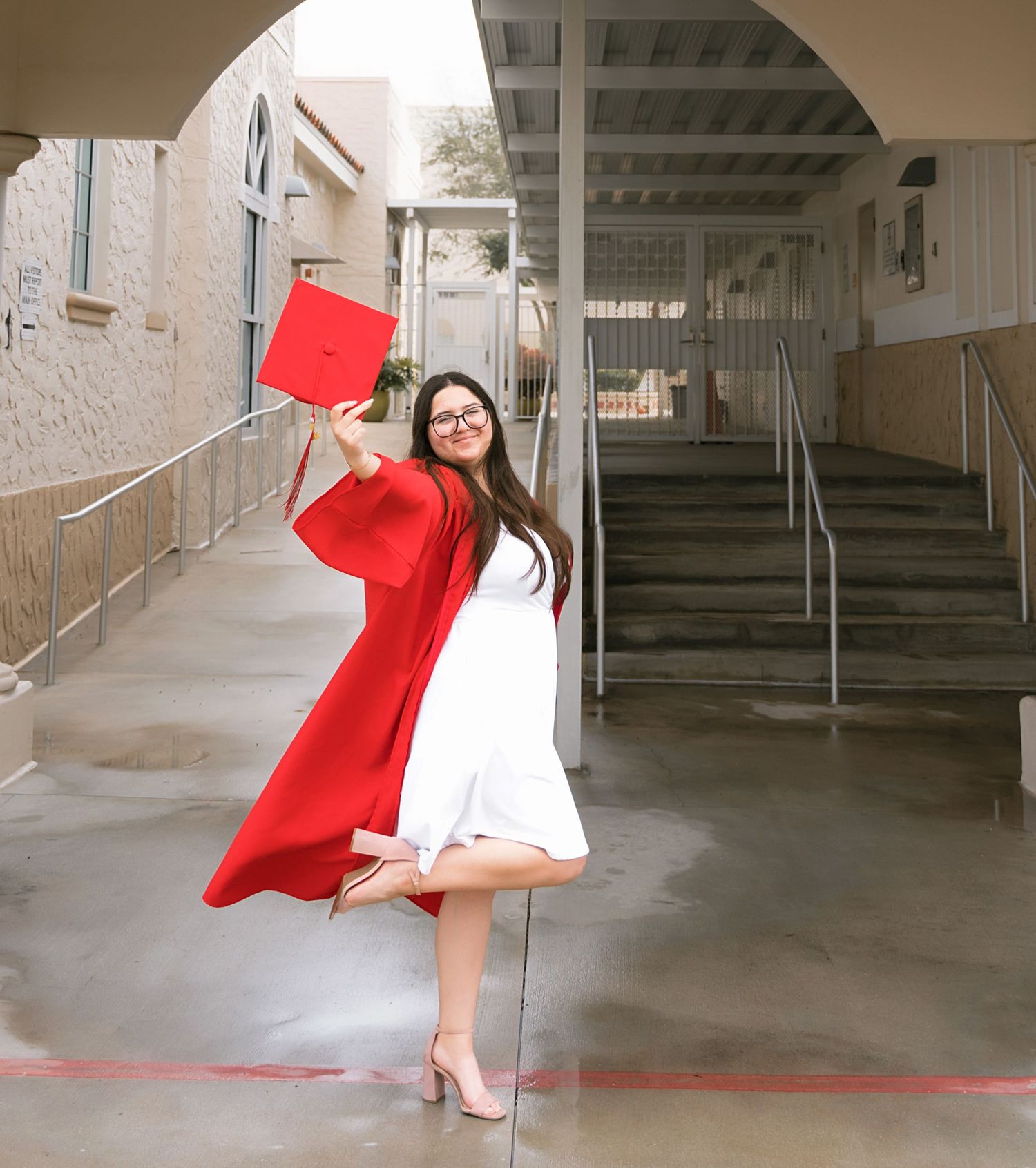 A graduate in white dress and red gown strikes a playful pose in a hallway.