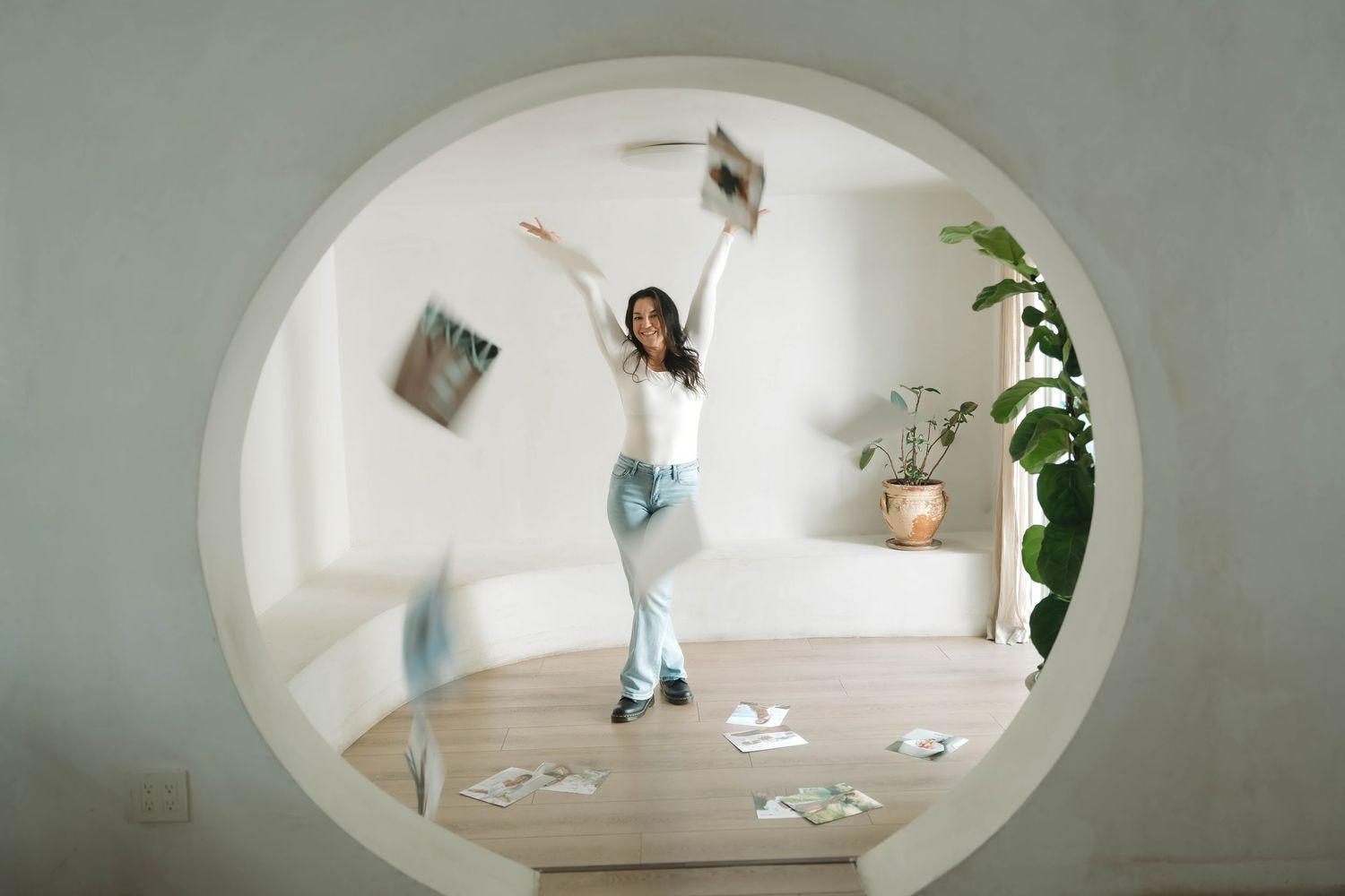 Modern white bathroom with oval window and minimalist decor featuring houseplants and natural light.