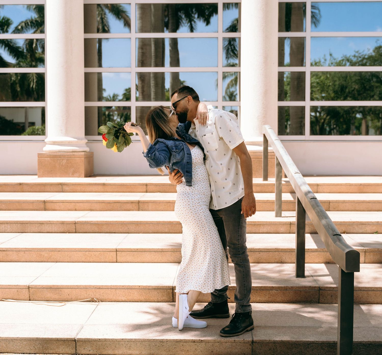 Romantic wedding couple embracing on outdoor stone steps with palm trees in background.