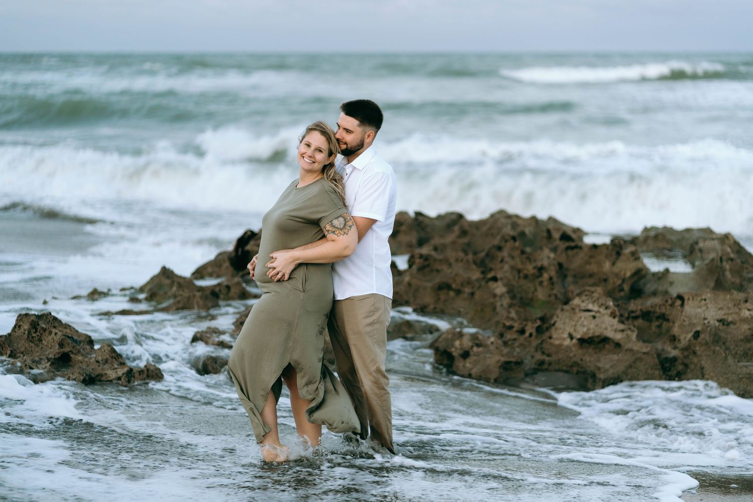 Couple embracing on rocky coastal shoreline with waves crashing in background.