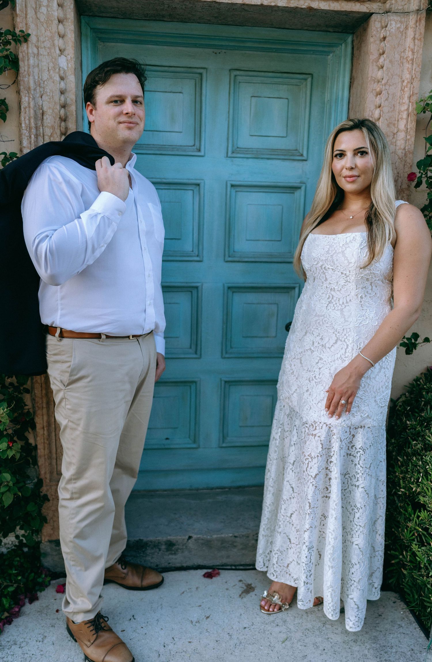 Couple posing in front of turquoise door, woman in white lace gown and man in white shirt with khaki pants.
