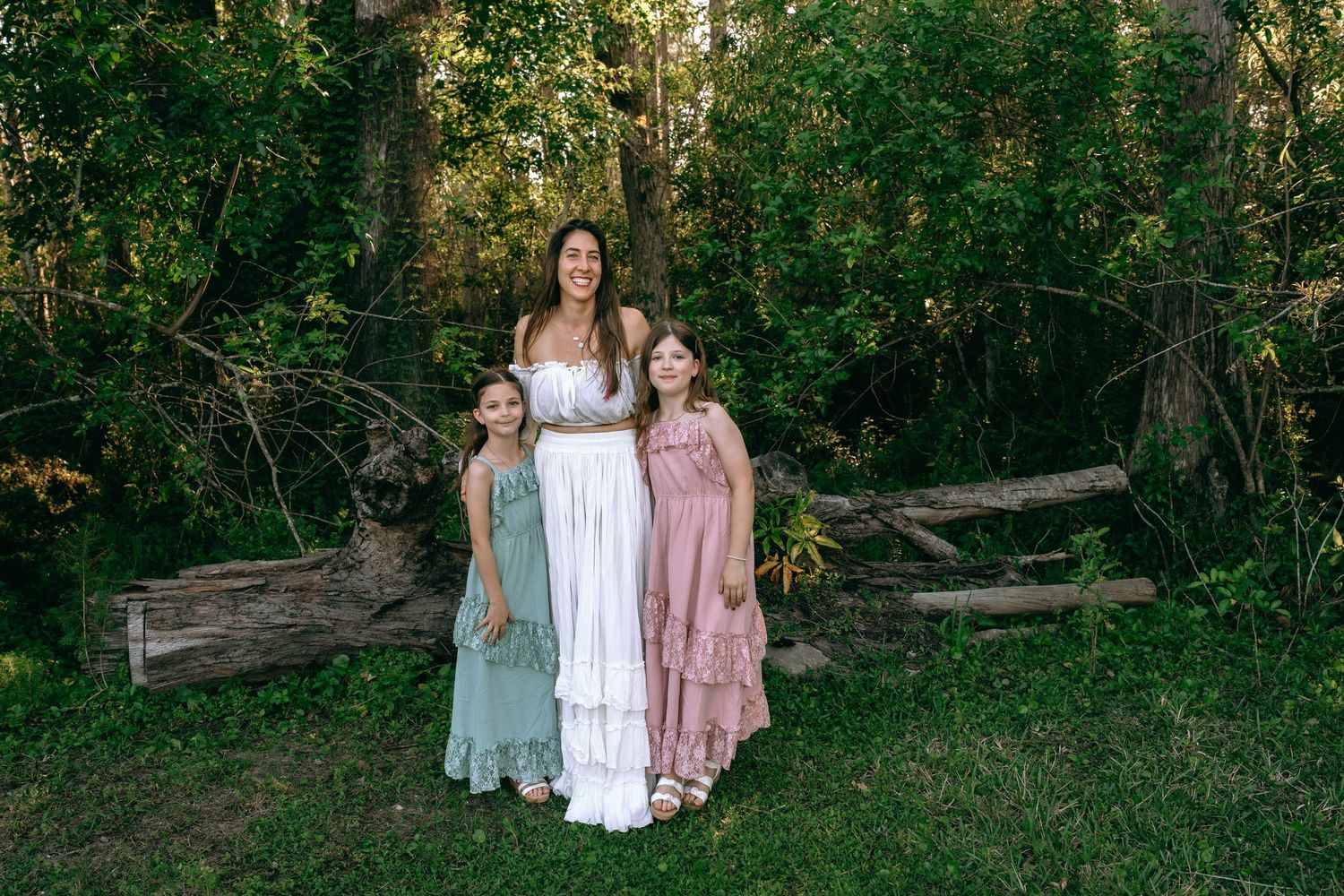 A mother in a white dress poses with two daughters in sage and pink dresses in a wooded setting. Colt Farms