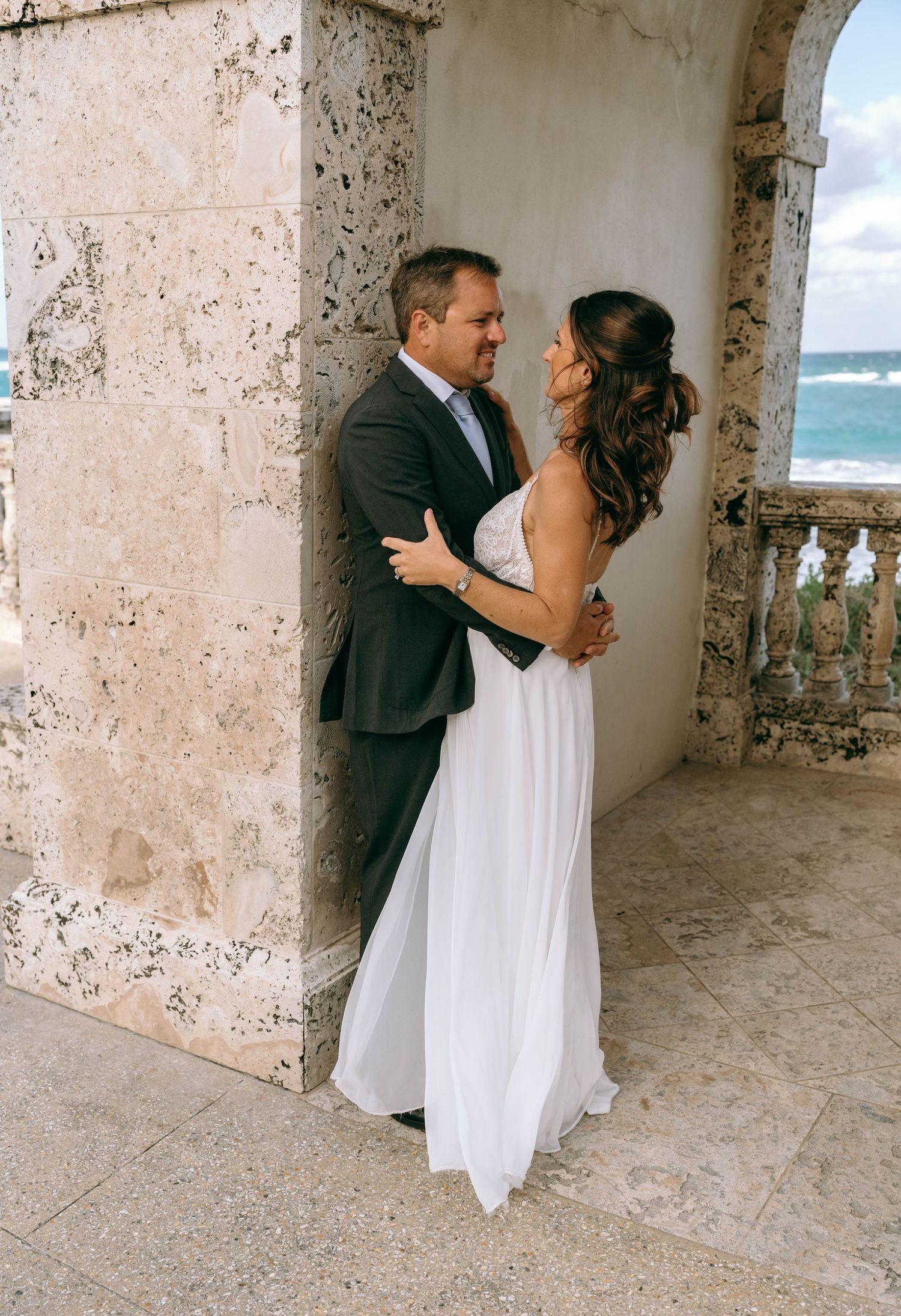 Bride and groom share an intimate embrace on a stone balcony overlooking the ocean.