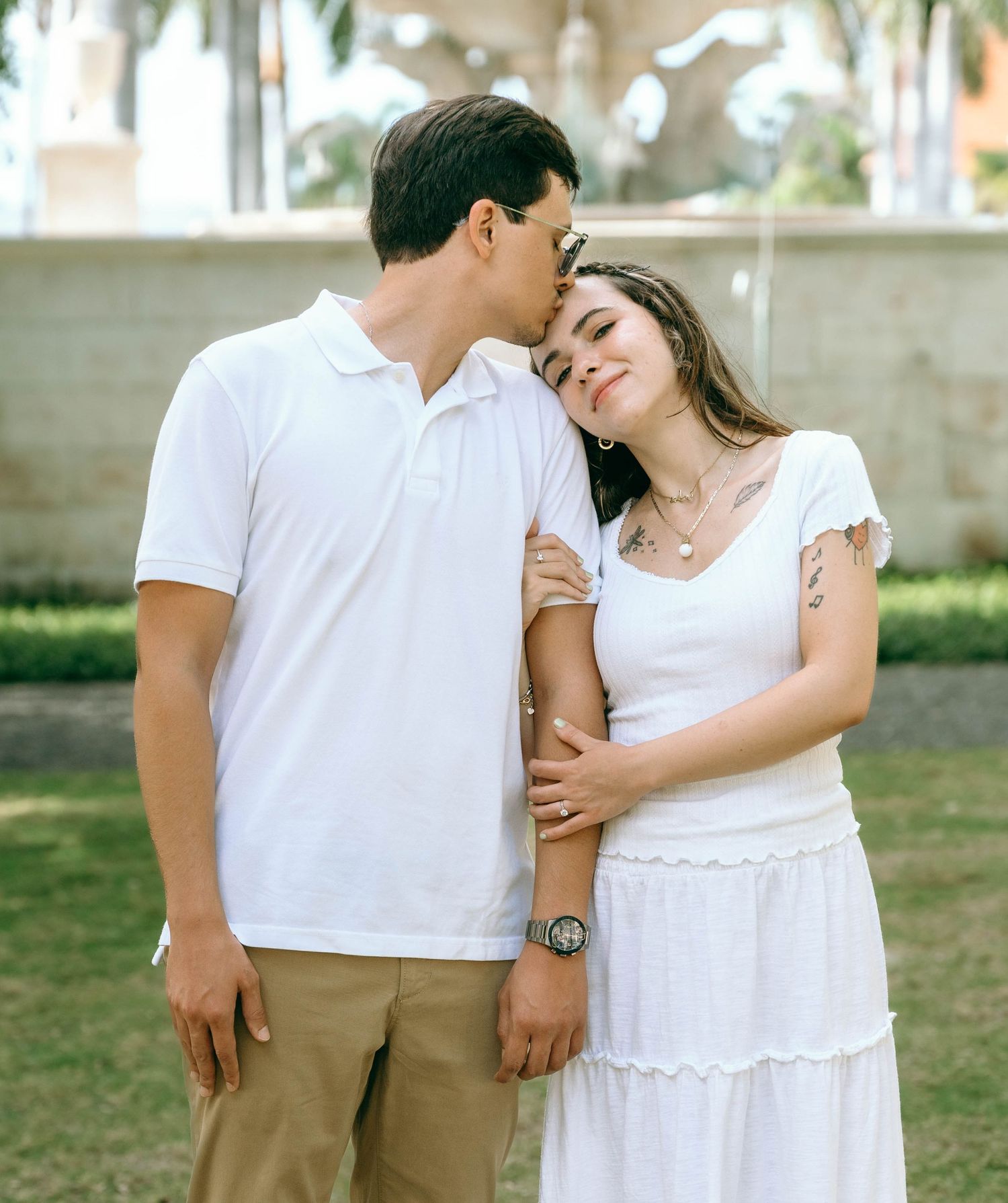 A couple in white outfits share a tender moment outdoors, with the man kissing the woman's forehead. Engagement photos on Worth Avenue