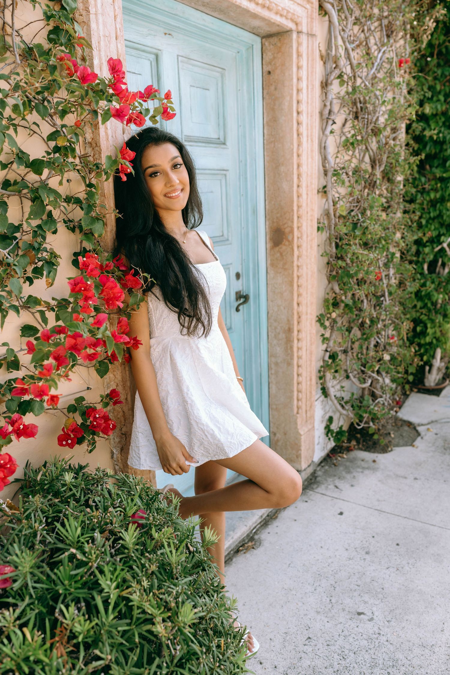 Young woman in white dress poses by blue door with red bougainvillea flowers and green vines on Worth Avenue Palm beach.