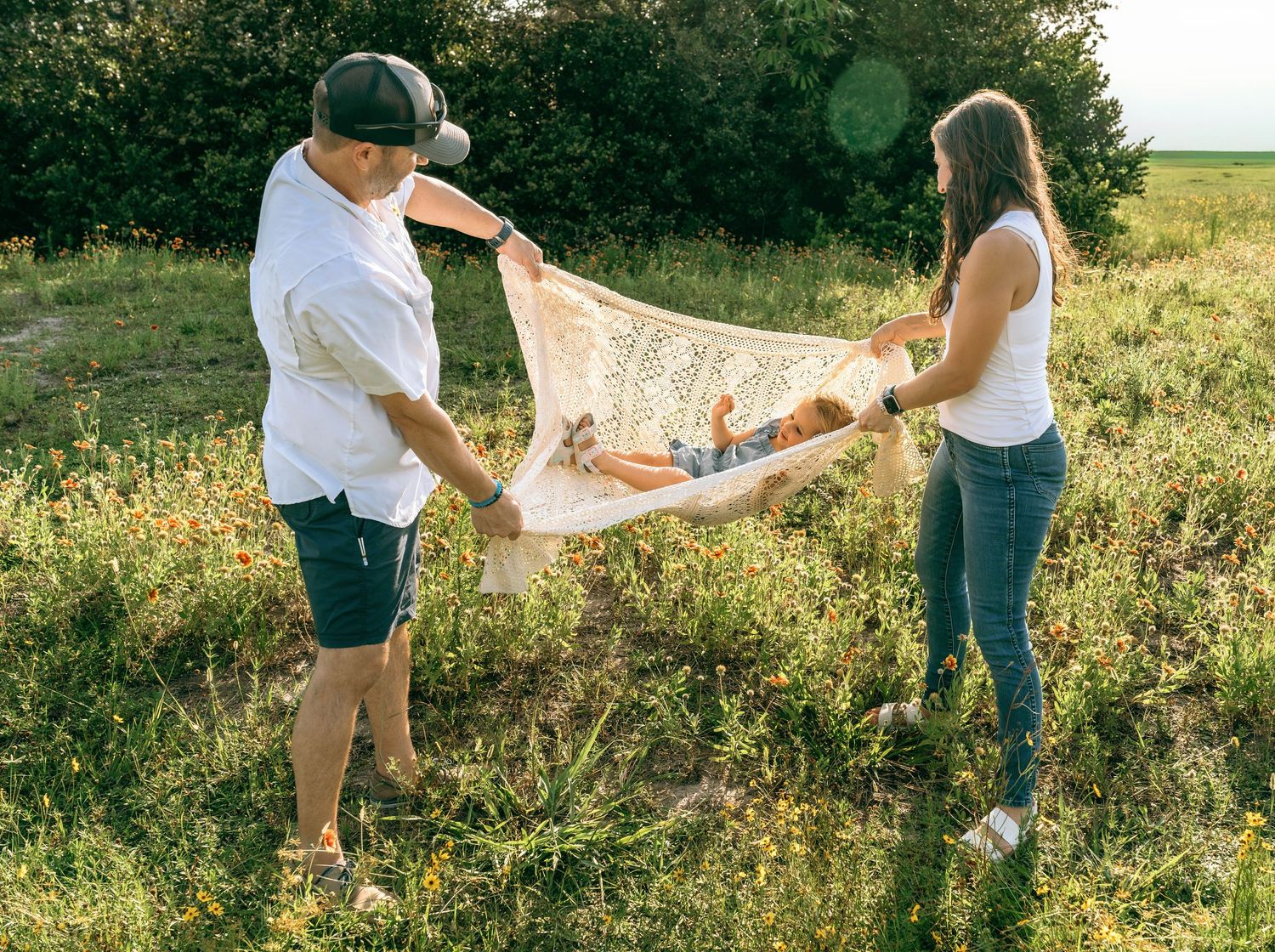 Two parents hold a baby in a blanket outdoors in a green field on a sunny day.