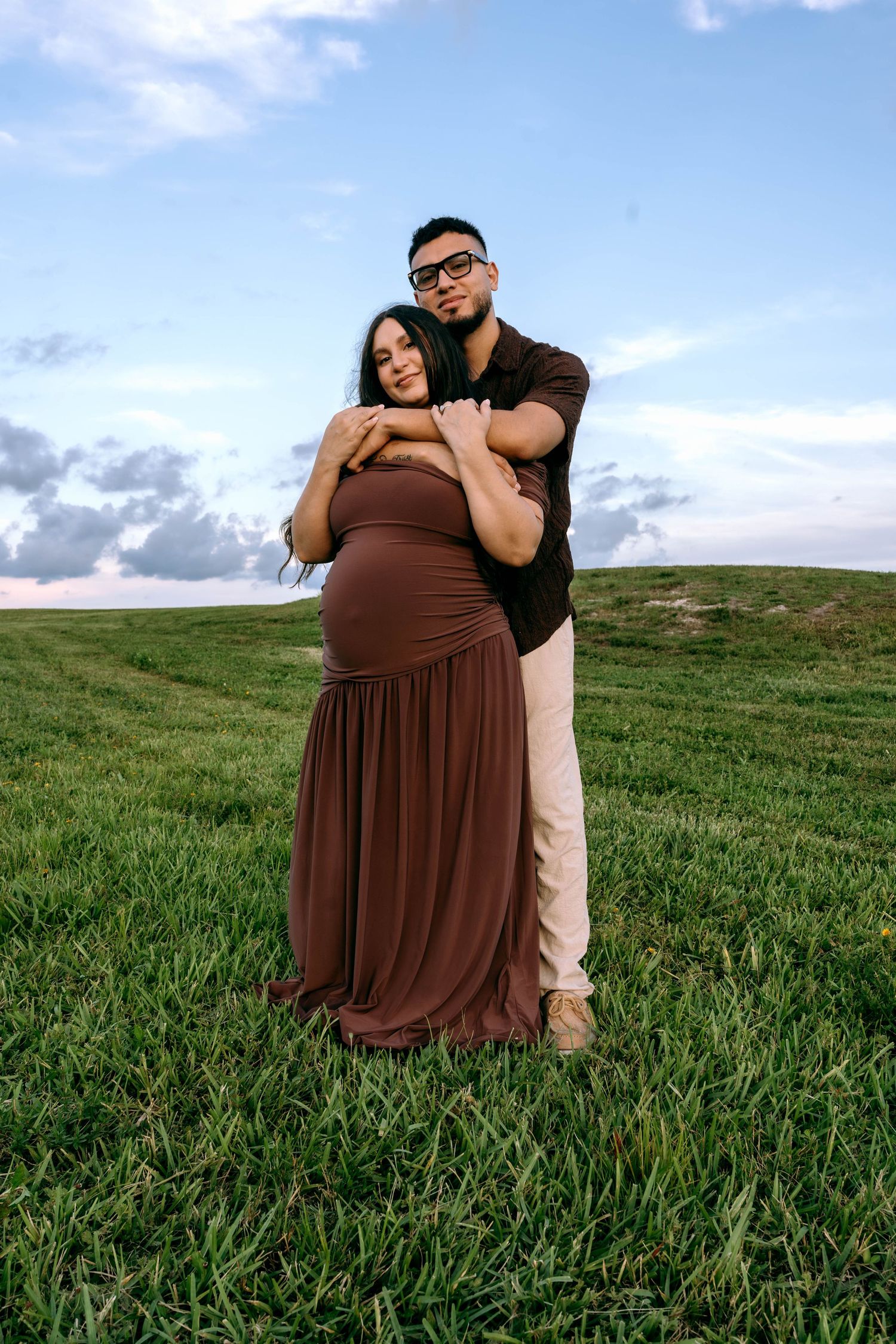 A pregnant woman in a brown dress embraced by her partner in a green field under a cloudy sky.