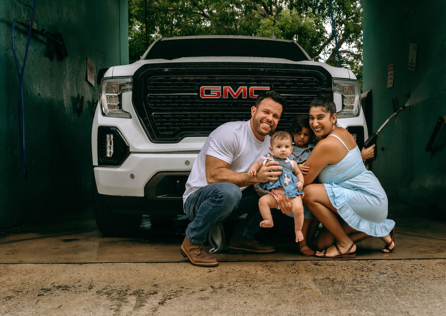 Happy family posing in front of a white GMC Sierra truck, parents holding baby in a garage setting.