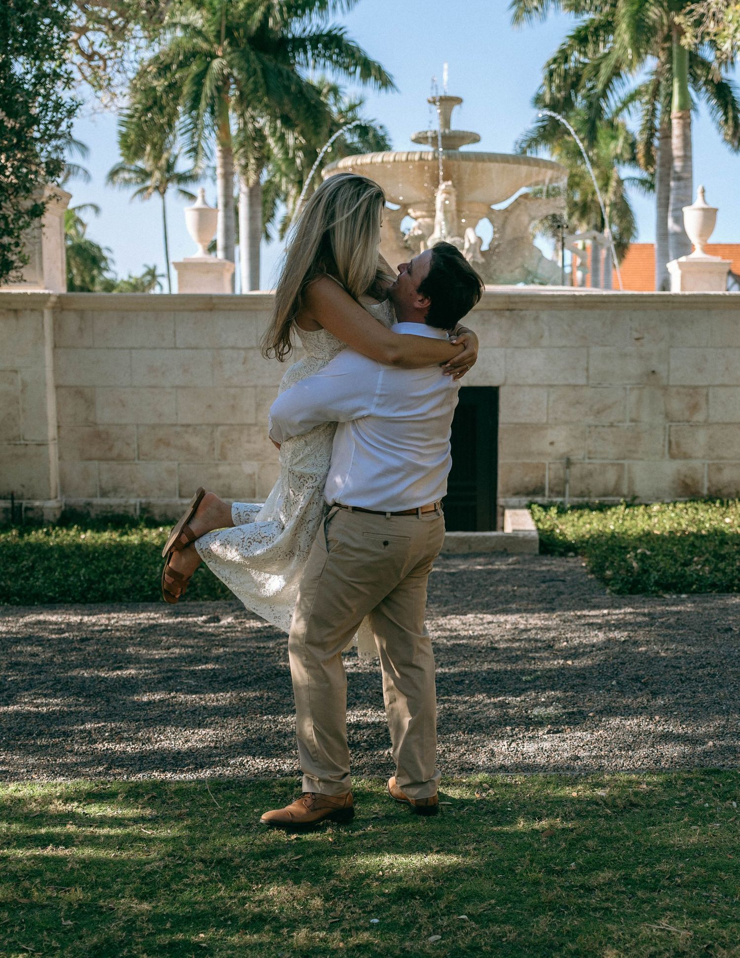 Couple sharing a romantic lift embrace in front of a fountain surrounded by palm trees.