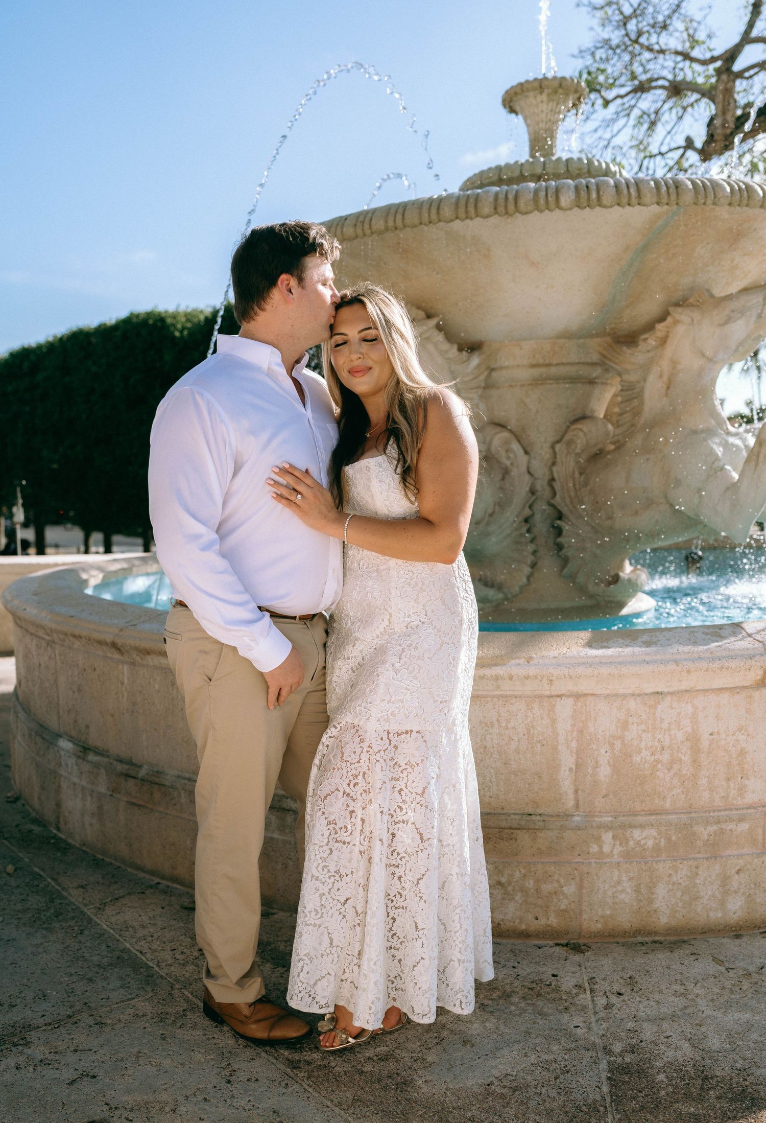Couple embracing near an ornate stone fountain outdoors on a sunny day, woman in lace dress.