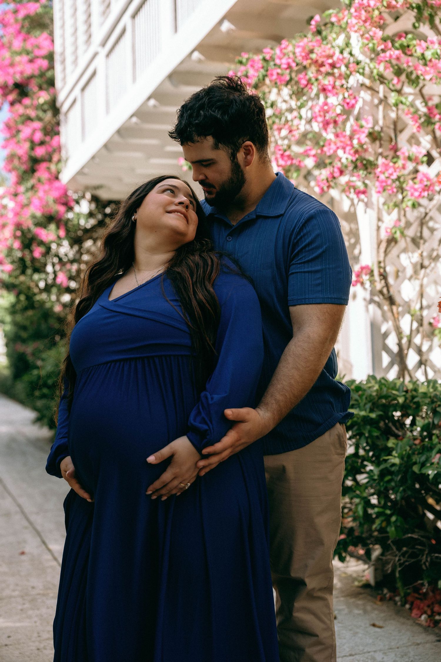 Couple sharing tender maternity moment outdoors, woman in blue dress with partner embracing her baby bump near pink flowers. Worth Avenue
