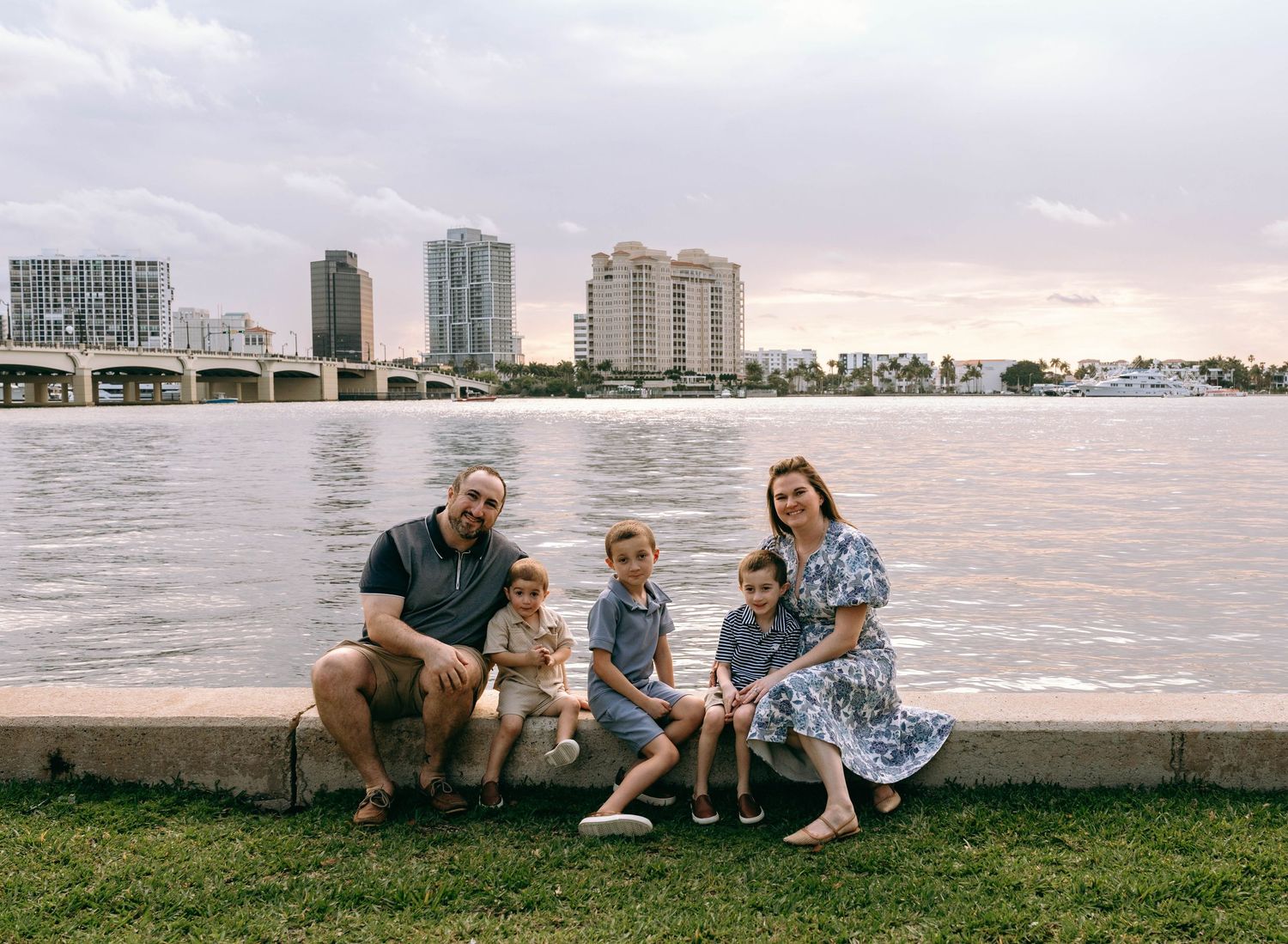 Family of five posing by waterfront with city skyline at sunset in Florida.  Family photos at Bradley Park Palm Beach, FL