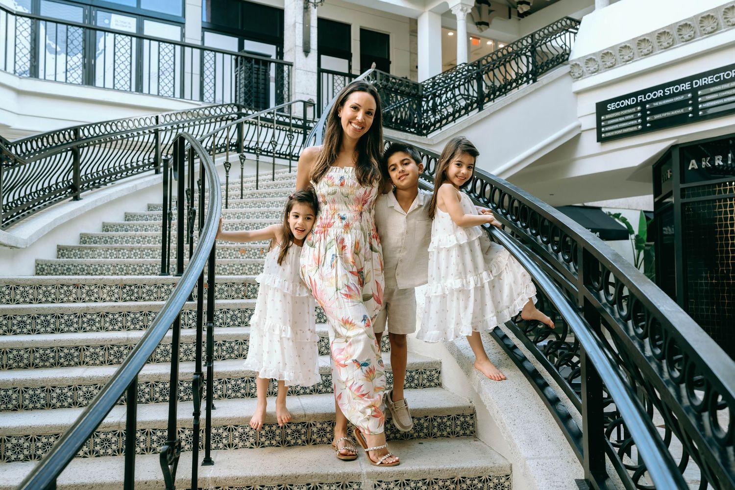 A woman in a floral jumpsuit poses on elegant stairs with three young girls in white dresses at a luxury hotel. Worth Avenue, Palm Beach, FL