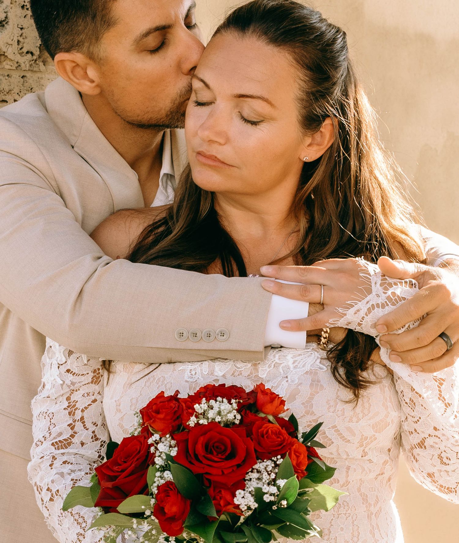Groom kissing bride on forehead while she holds a bouquet of red roses, wearing a white lace wedding dress.