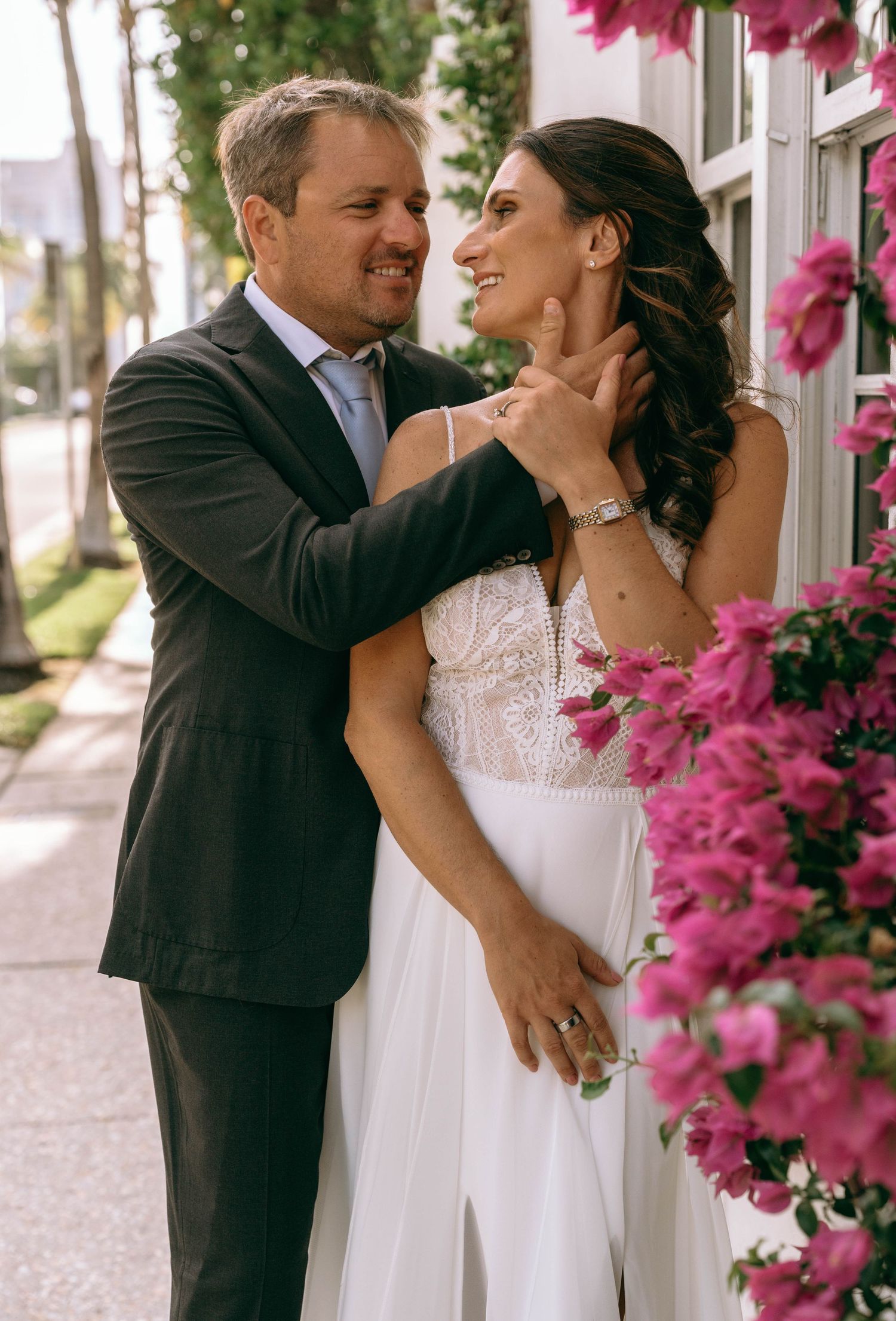 Groom tenderly holds brides face as they smile at each other near pink bougainvillea blooms.