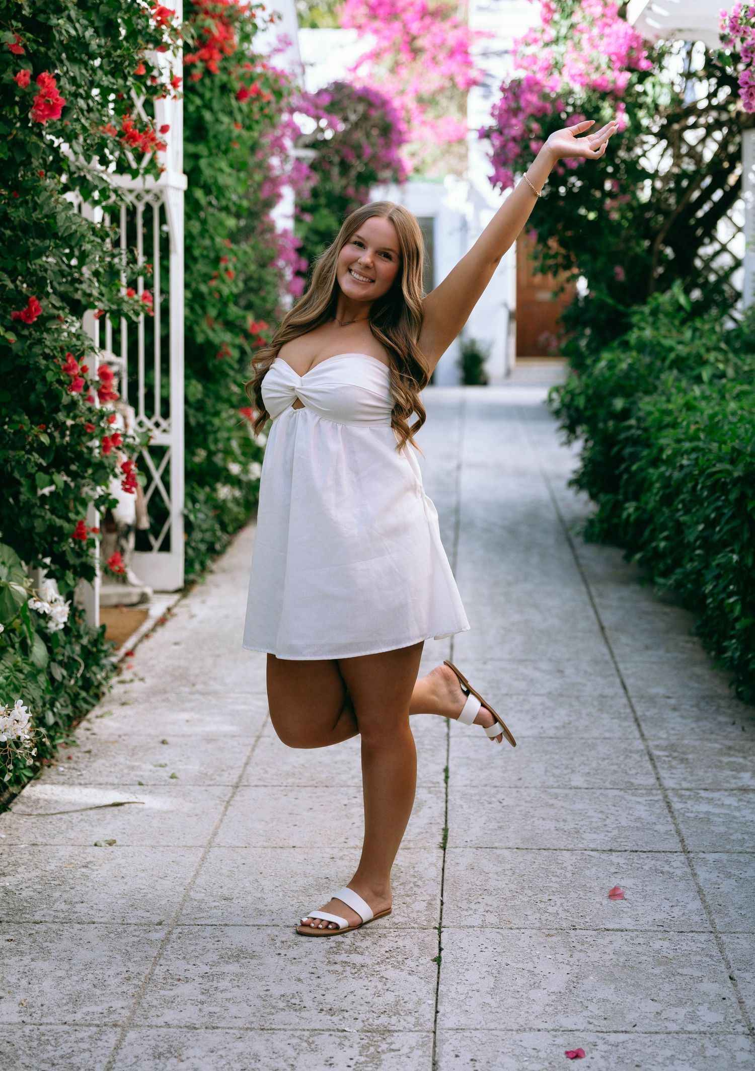 Young woman in white mini dress posing joyfully in a flower-lined Mediterranean walkway.