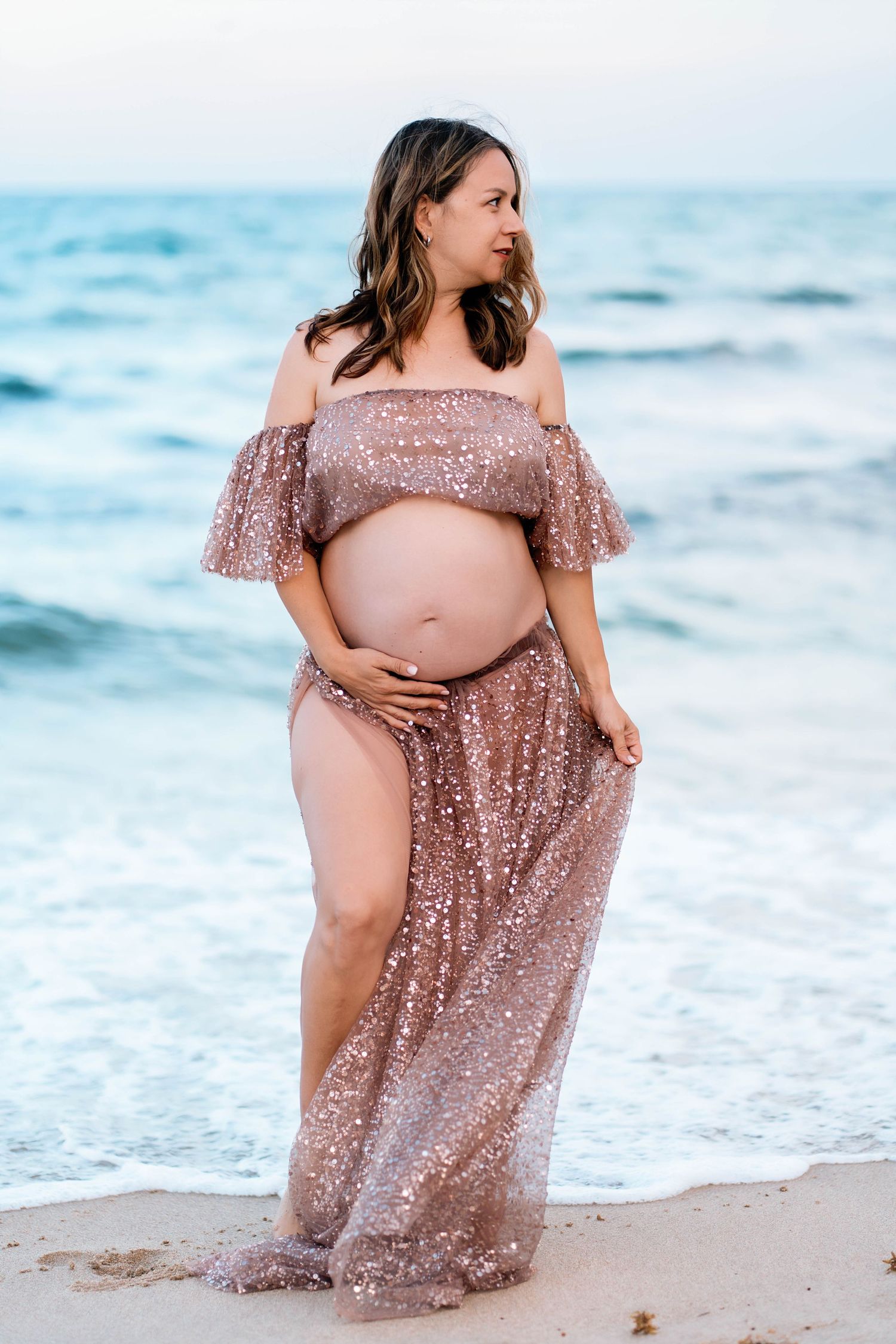 Maternity beach photos with a sparkly rose gold dress against ocean waves.
