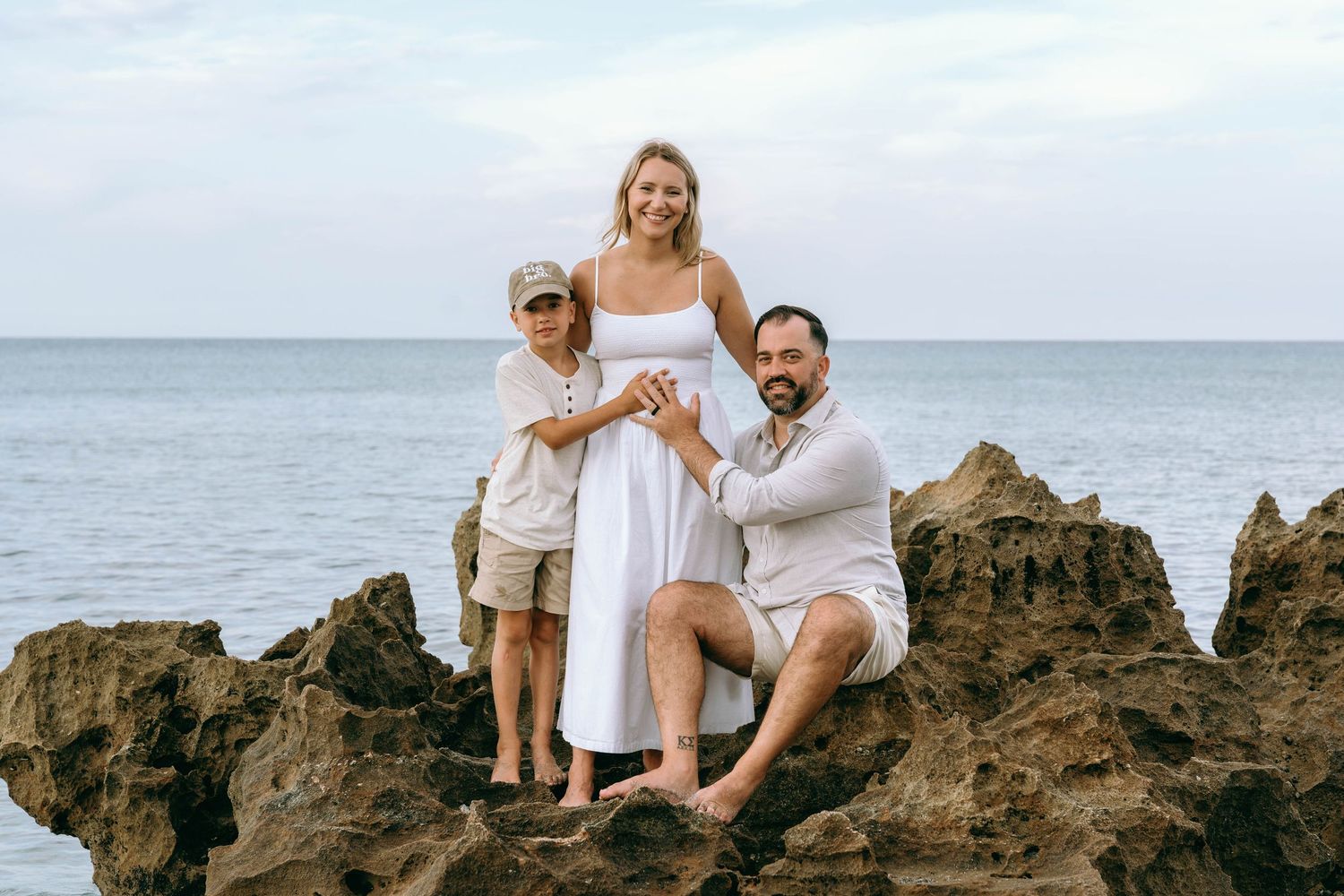 Family of three posing on rocky beach, pregnant woman in white dress with husband and son, ocean backdrop.