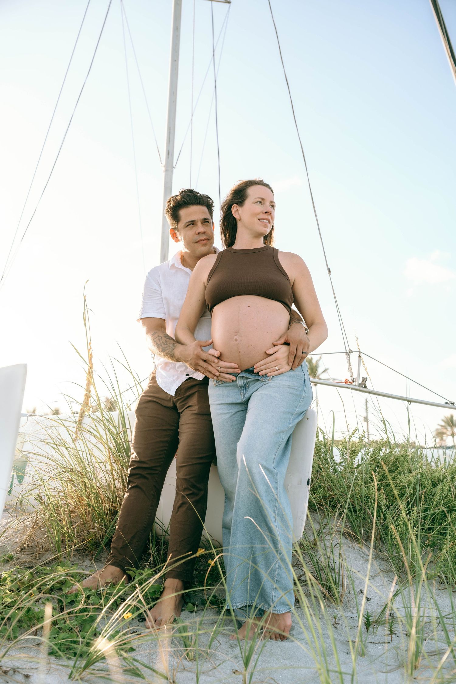 Pregnant couple standing on sandy beach dunes near sailboat masts, woman in brown crop top and jeans with partner embracing her bump.