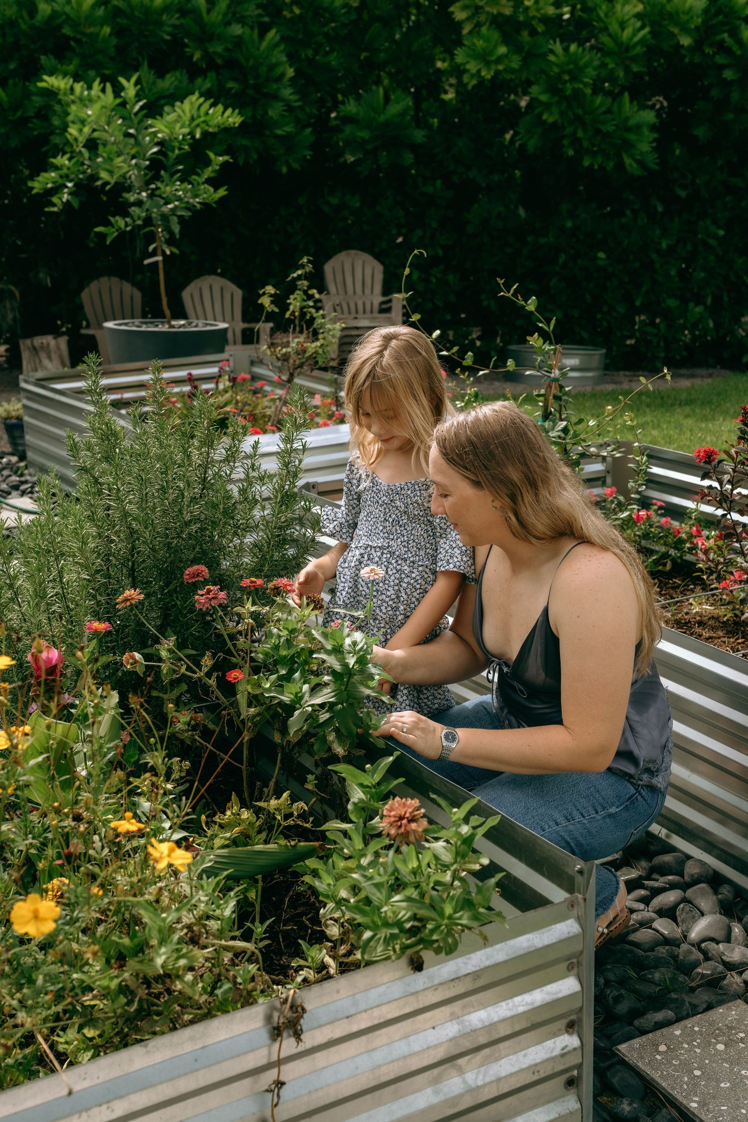 A woman and young girl examine colorful flowers in raised metal garden beds surrounded by lush greenery.