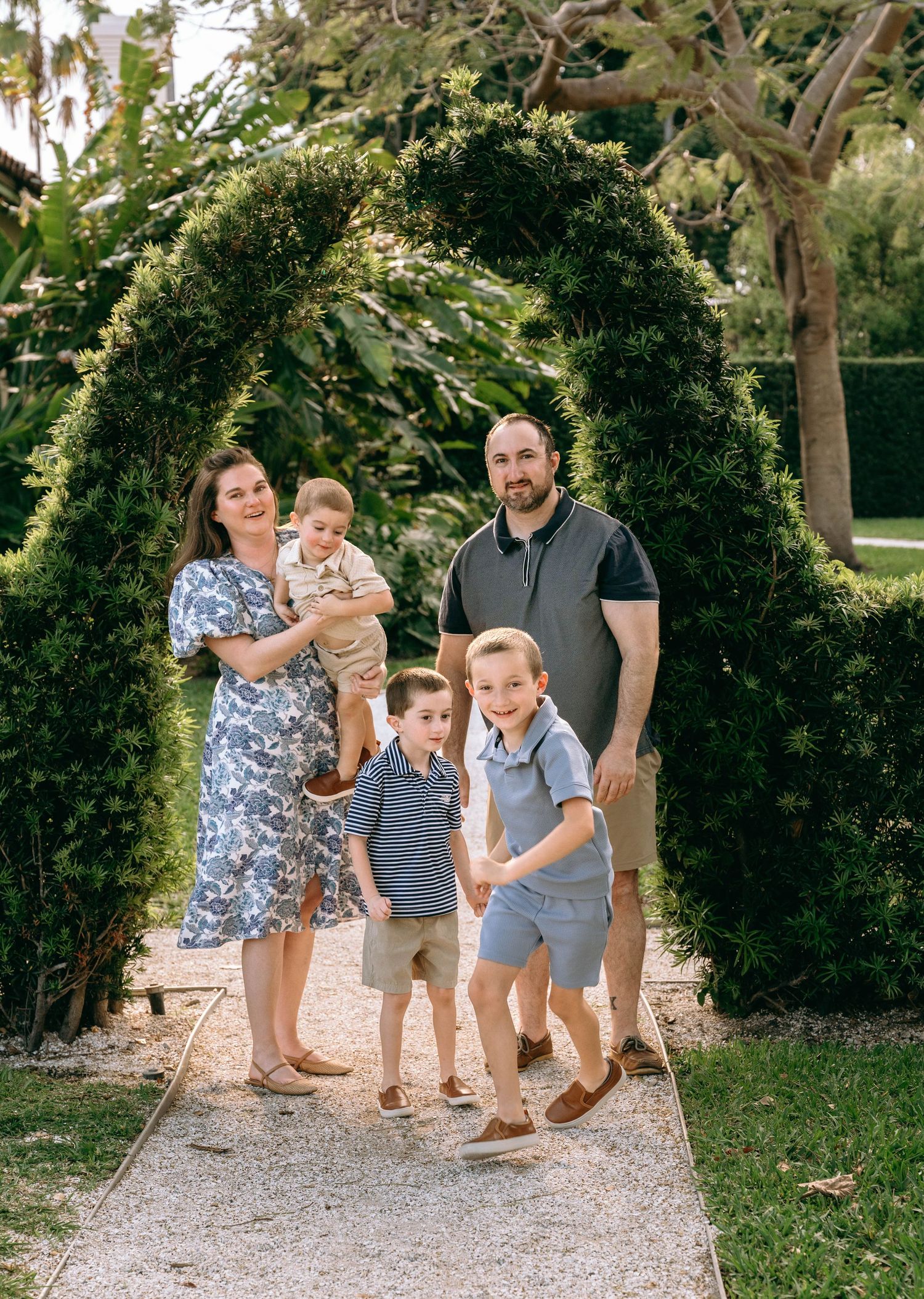 Family of five posing under a lush green arch on a garden path, parents with three young boys in coordinating outfits at Bradley Park in Pa.