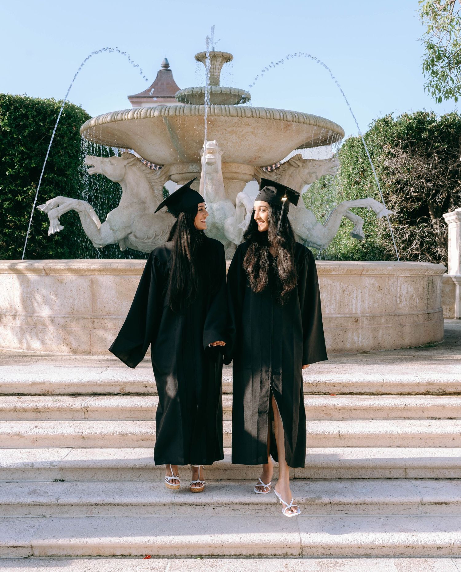 Two female graduates in black caps and gowns pose back-to-back before an ornate stone fountain on Worth Avenue Palm beach.