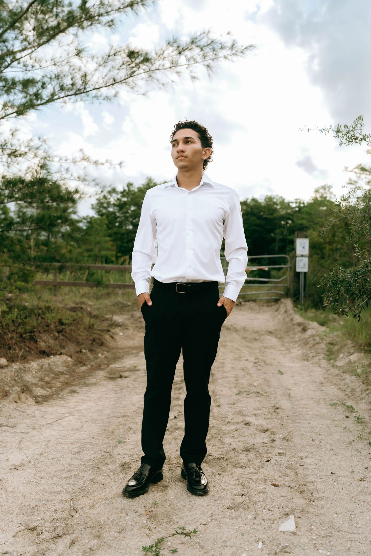 Young man in white dress shirt and black pants stands on dirt path looking up at cloudy sky. Senior Photos at Colt Farms in Boynton Beach