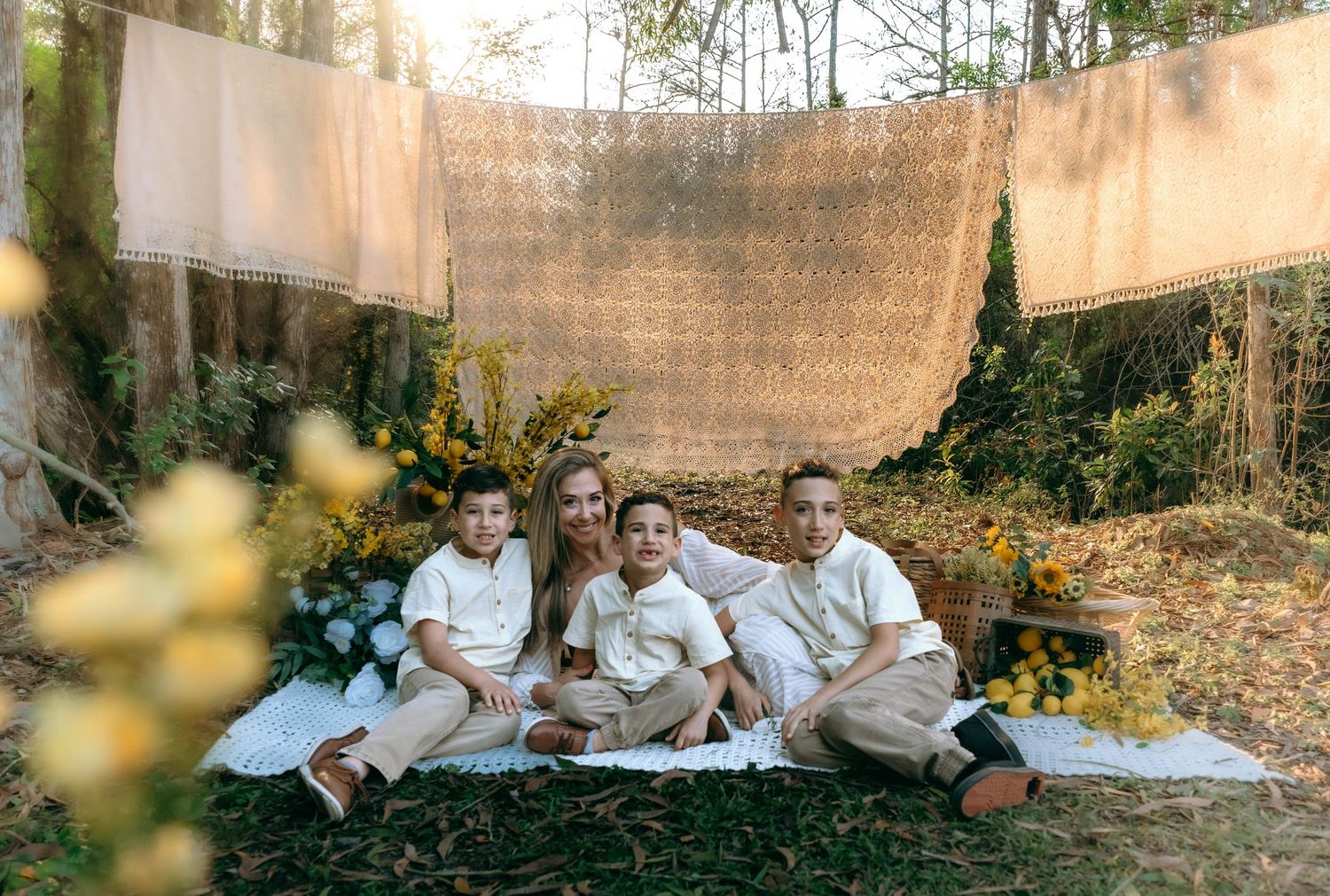 A family wearing white outfits sits together on blankets during an outdoor photo session in a natural setting.