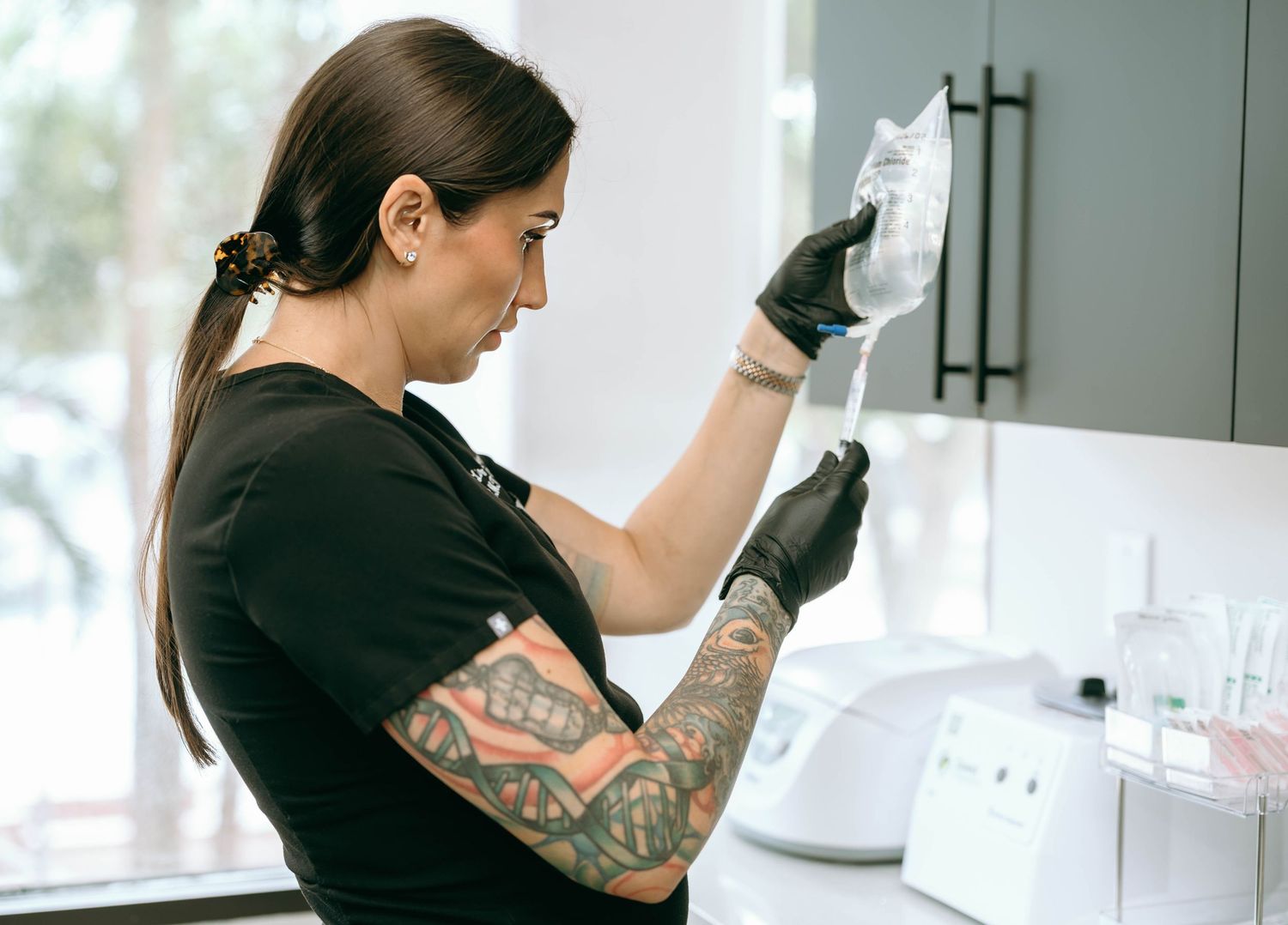 Tattooed female healthcare professional in black gloves examining an IV bag in a medical clinic.