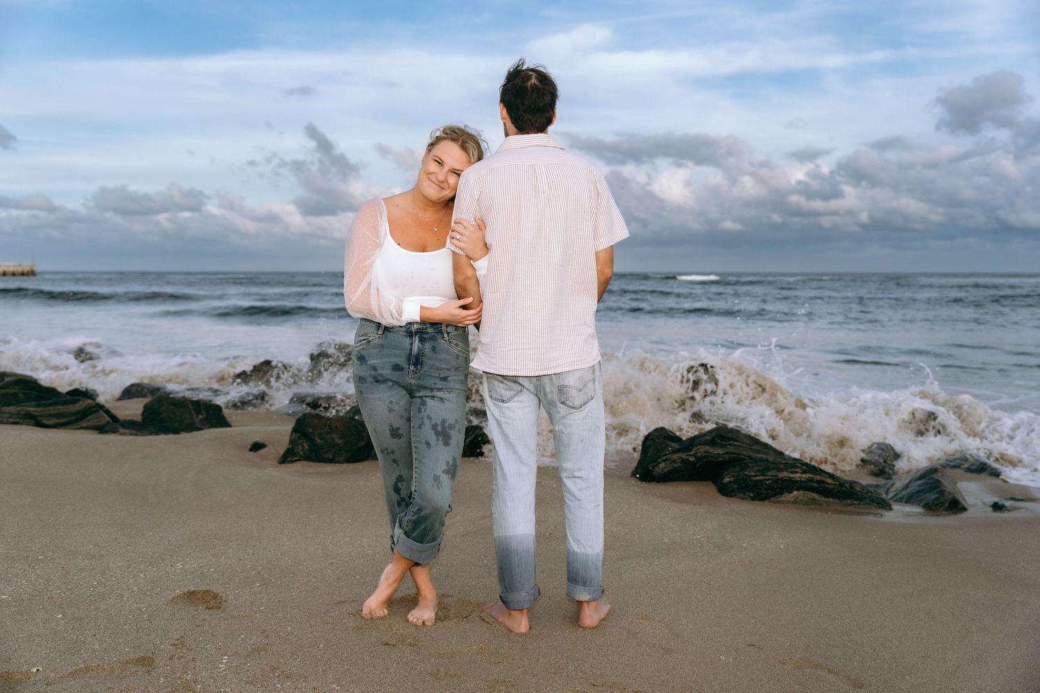 A couple stands together watching the ocean at sunset with rocky coastline visible.