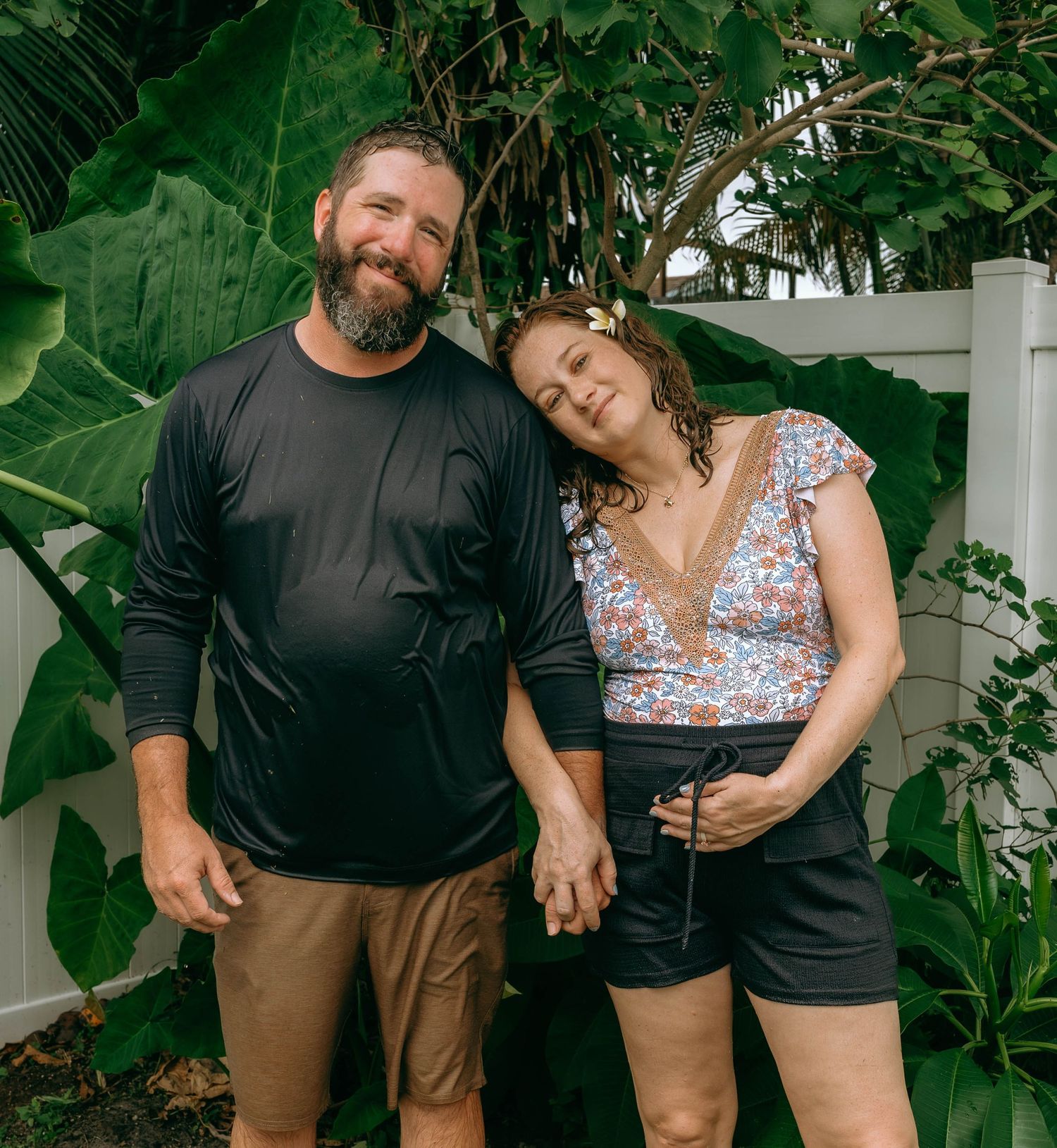 A couple holds hands and smiles in a lush tropical garden, the woman resting her head on the mans shoulder.