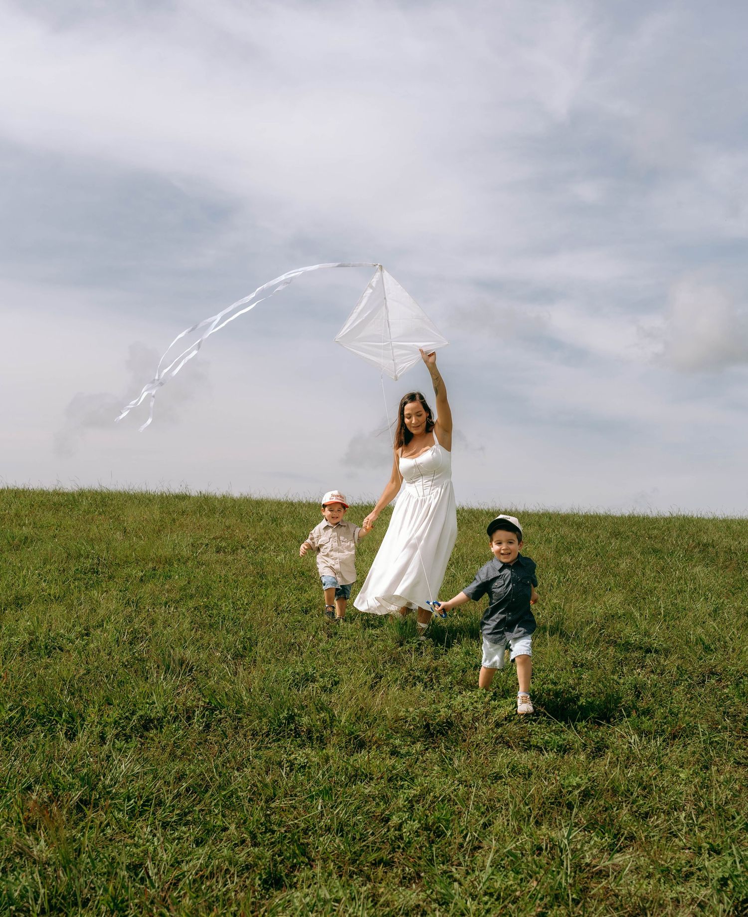 A smiling mother in a white dress holds a white kite while running with two children on a grassy hill.
