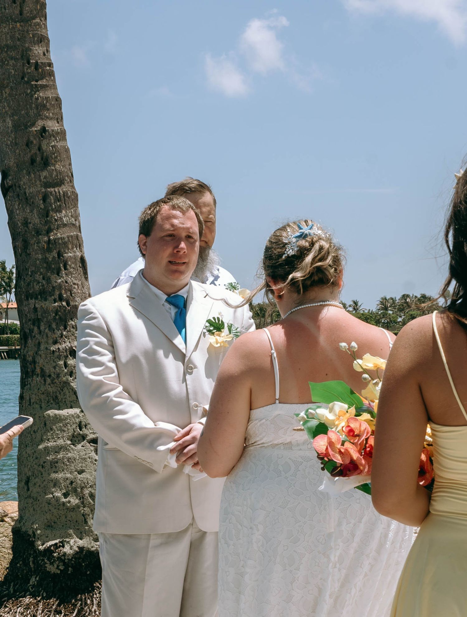 Groom in white suit exchanges vows with bride holding colorful bouquet at outdoor beach wedding ceremony.