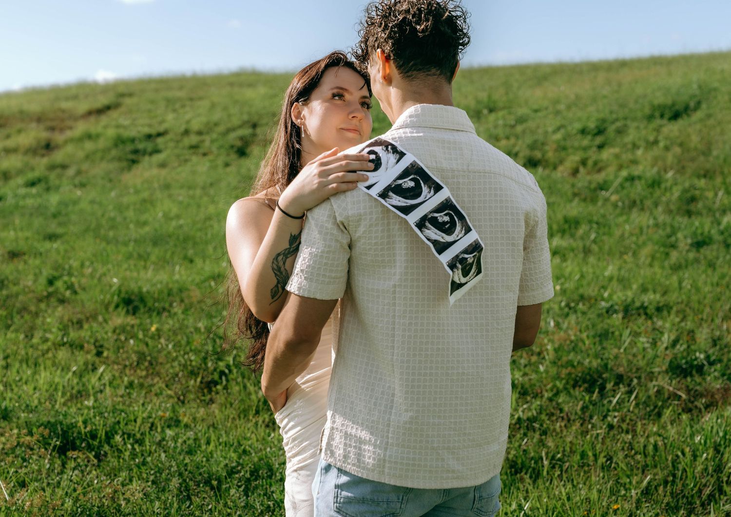 Couple embracing on a green grassy hill, woman touching mans face while he holds ultrasound photos.