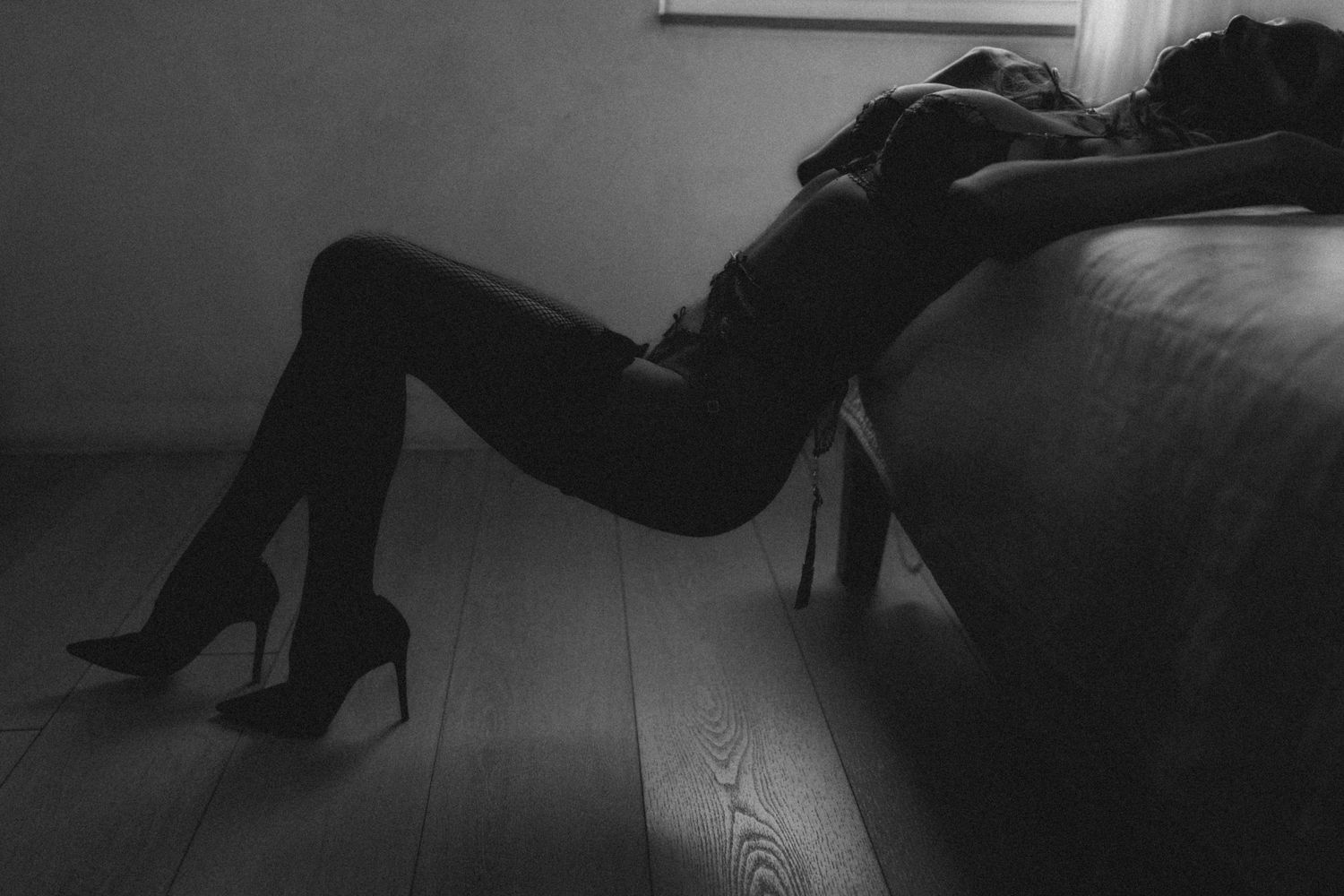 Black and white photo of a woman in high heels reclining against a bed on a wooden floor.