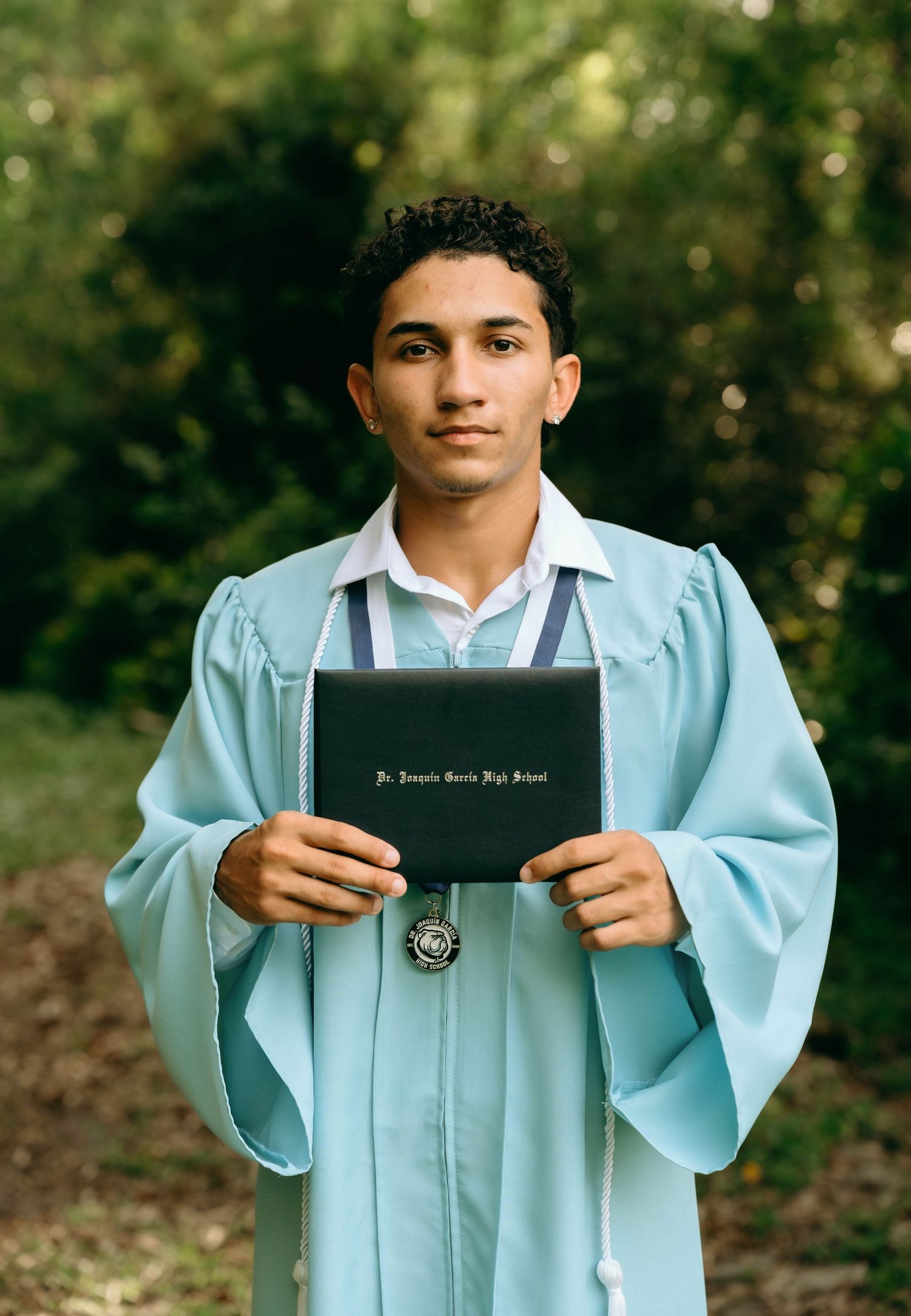 Young man in light blue graduation gown holding diploma from Joaquin Varela High School outdoors. Senior Photos at Colt Farms in Boynton