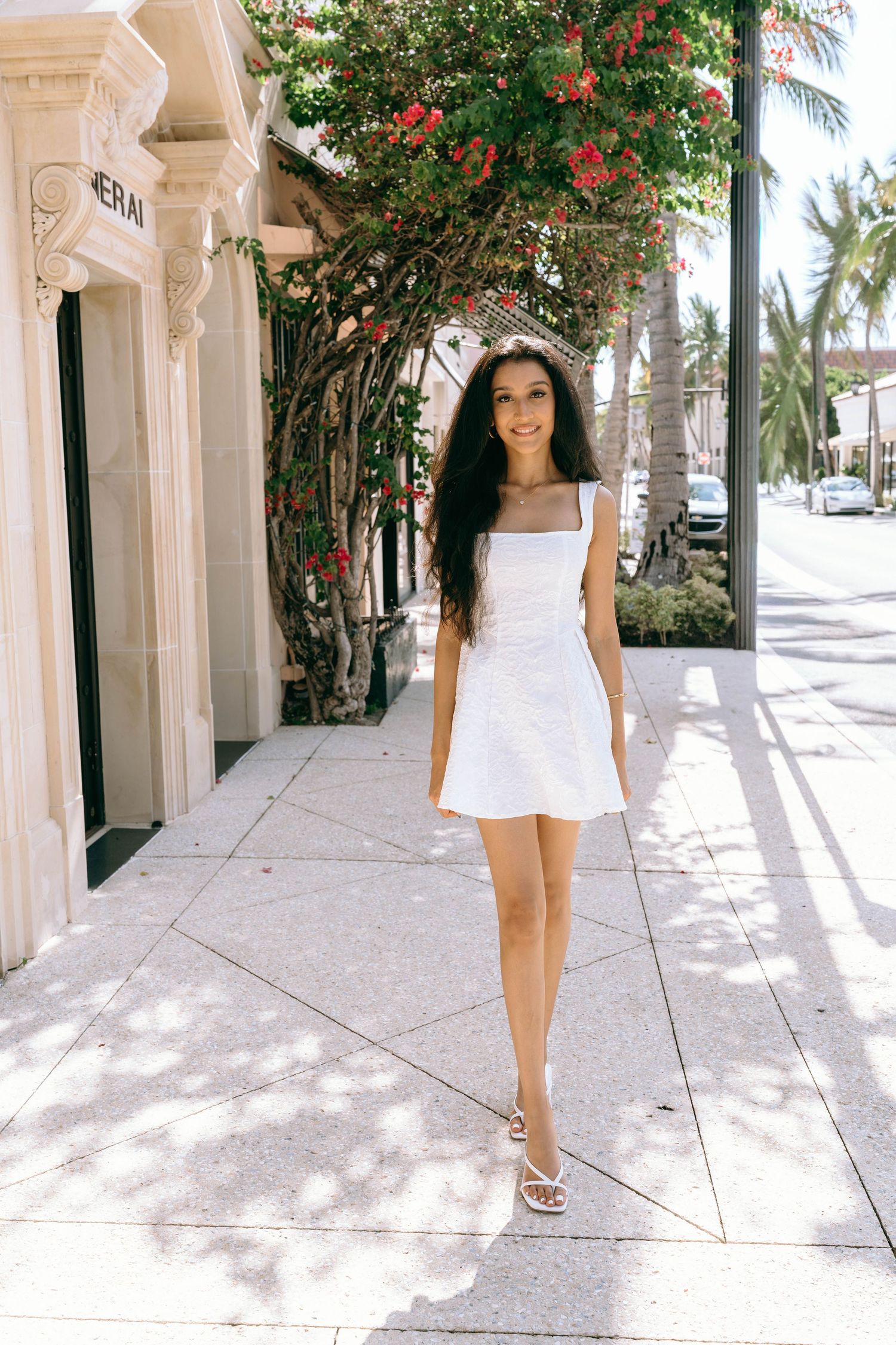 Young woman in white sundress walks sunny sidewalk lined with red bougainvillea and palm trees on Worth Avenue Palm beach.