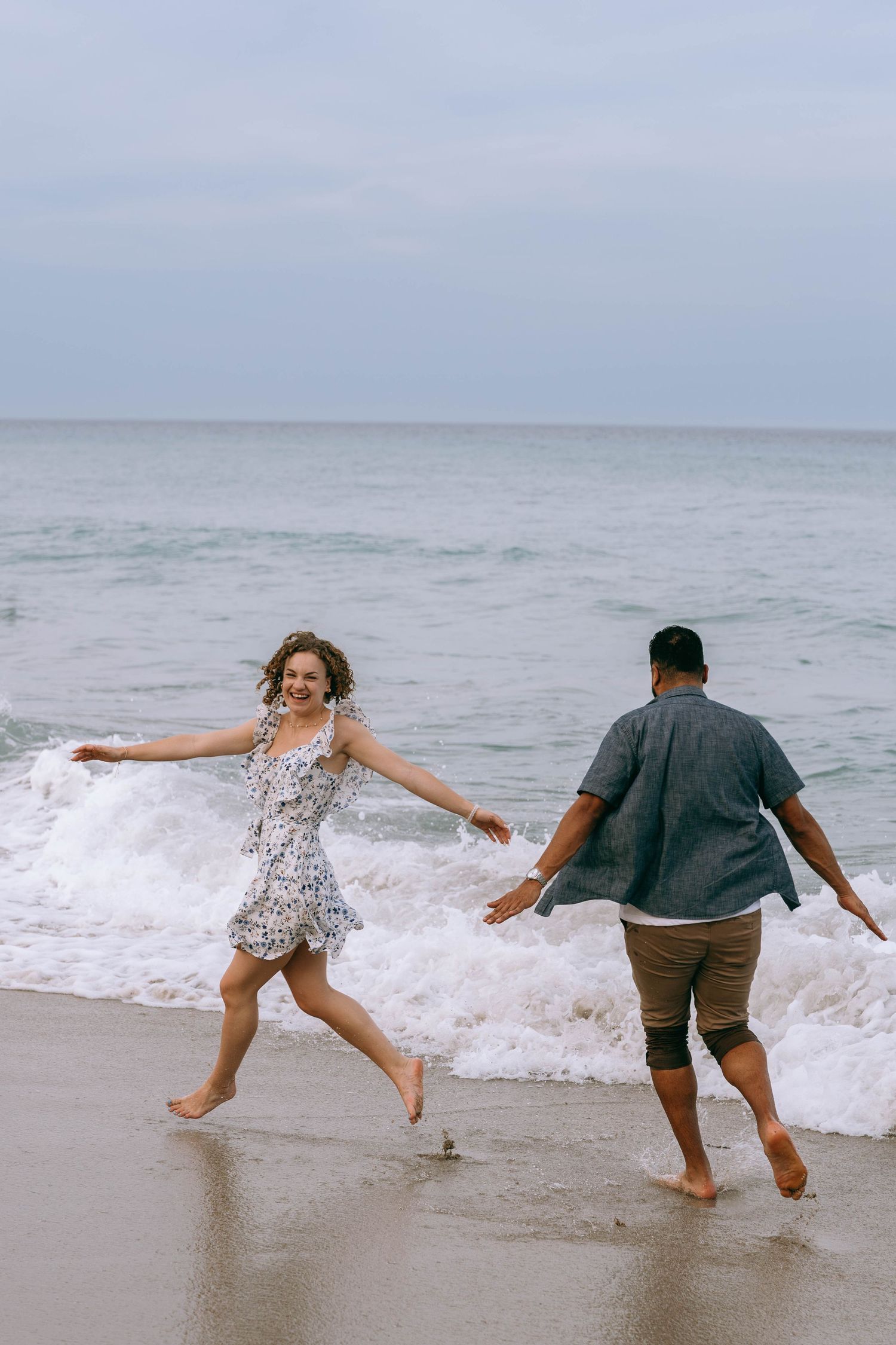Playful couple running barefoot along ocean shoreline, laughing as waves wash up on the beach on Worth Avenue Palm beach.